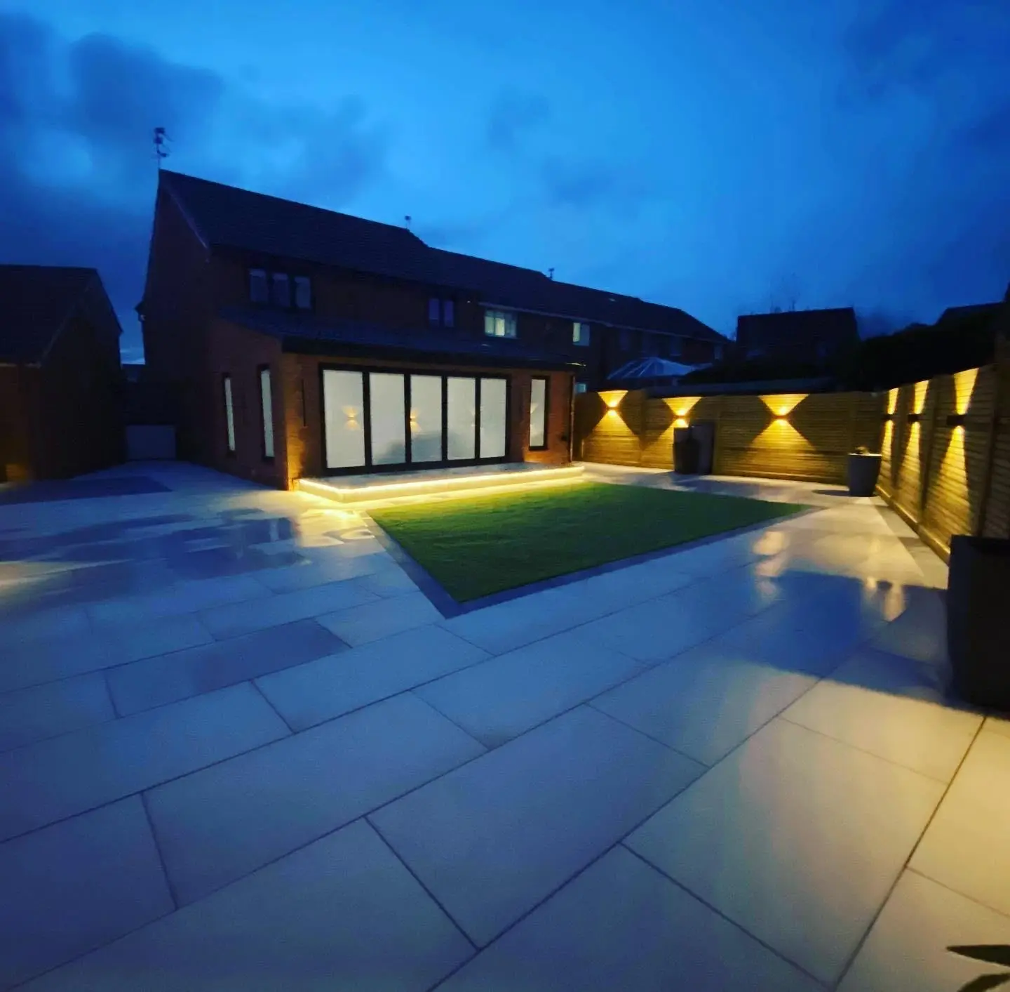 A rear garden at dusk with LED tape lighting under the raised patio step and up and down wall lights along the slatted fence.
