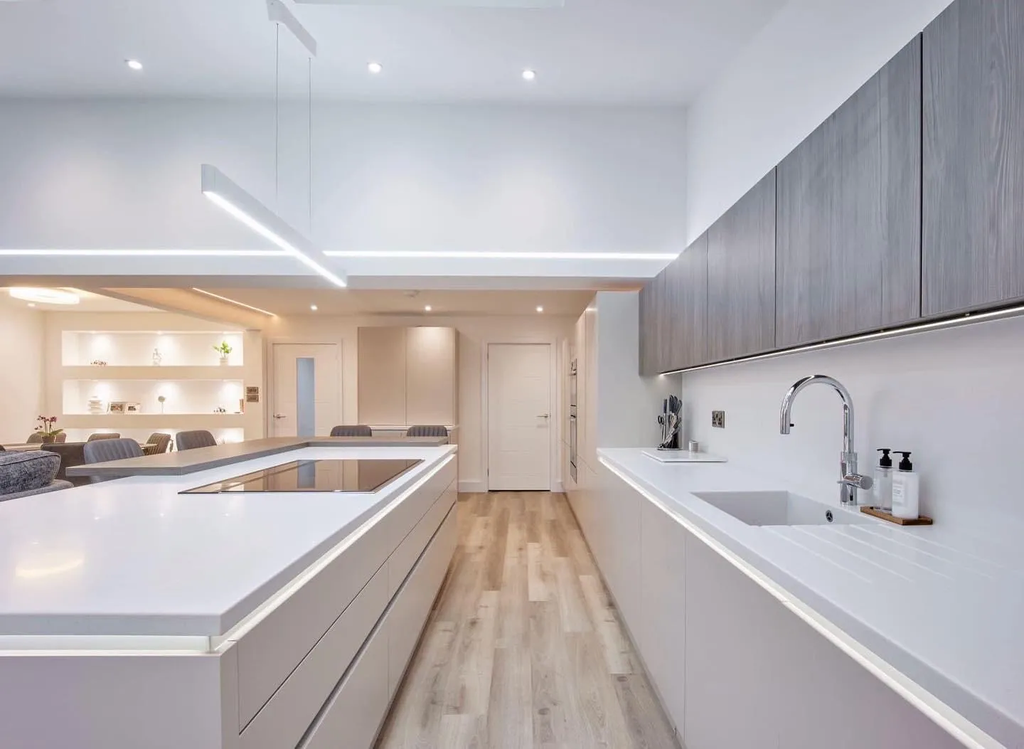A handleless kitchen with a linear LED pendant over the island hob, an LED profile along the ceiling bulkhead, downlights, and LED tape under the wall units and along the worktop edges.