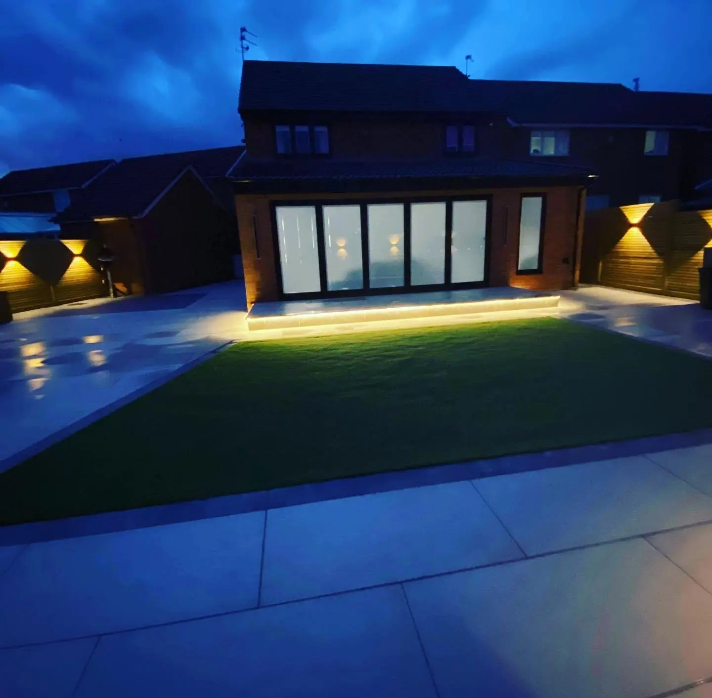 A rear garden at dusk looking back at the house, with LED tape under the patio step and up and down lights along the fences on both sides.