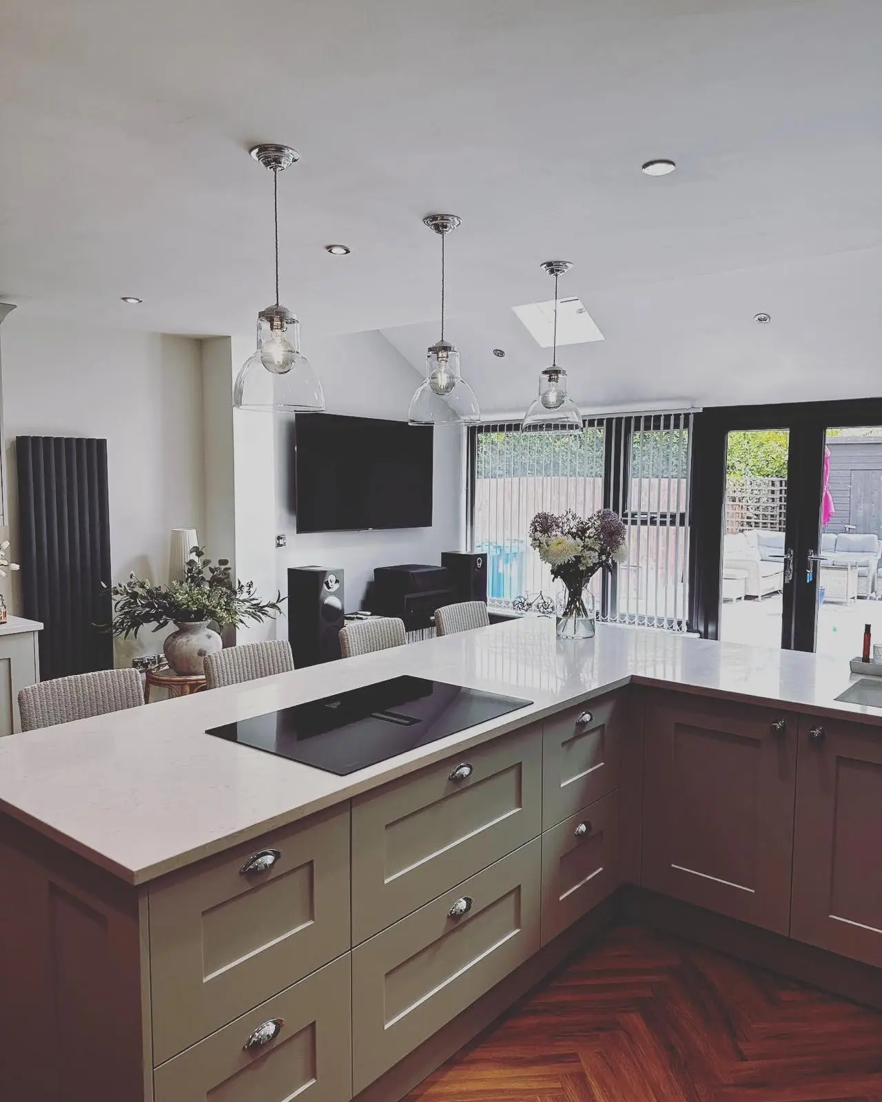 Three clear glass pendants hung over a peninsula with a hob, with LED downlights across the ceiling and a wall-mounted TV beyond.