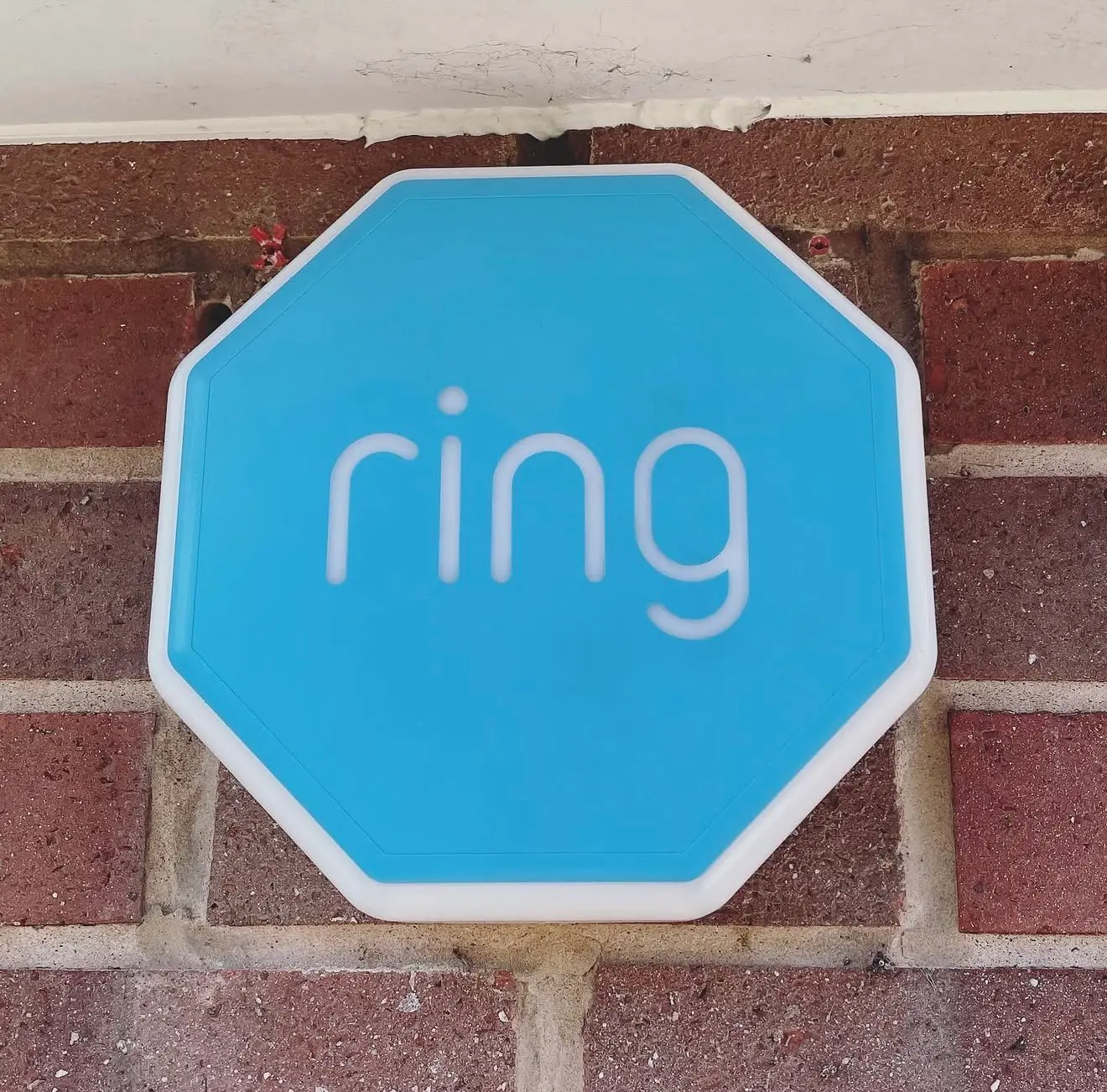 A blue Ring alarm siren fitted high on a red brick wall just below the white soffit.