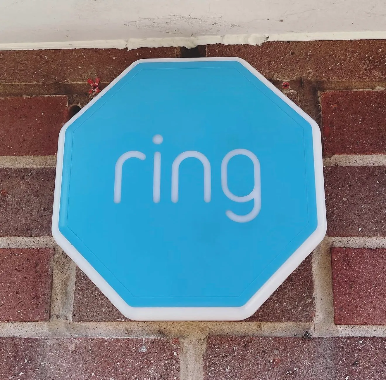 A blue Ring alarm siren fitted high on a red brick wall just below the white soffit.