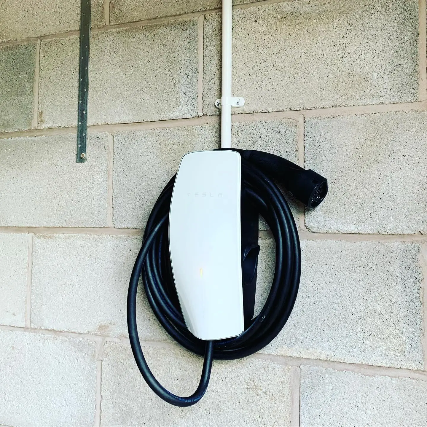 A white Tesla wall connector fixed to blockwork, with its cable coiled around it and the supply run in white conduit from above.