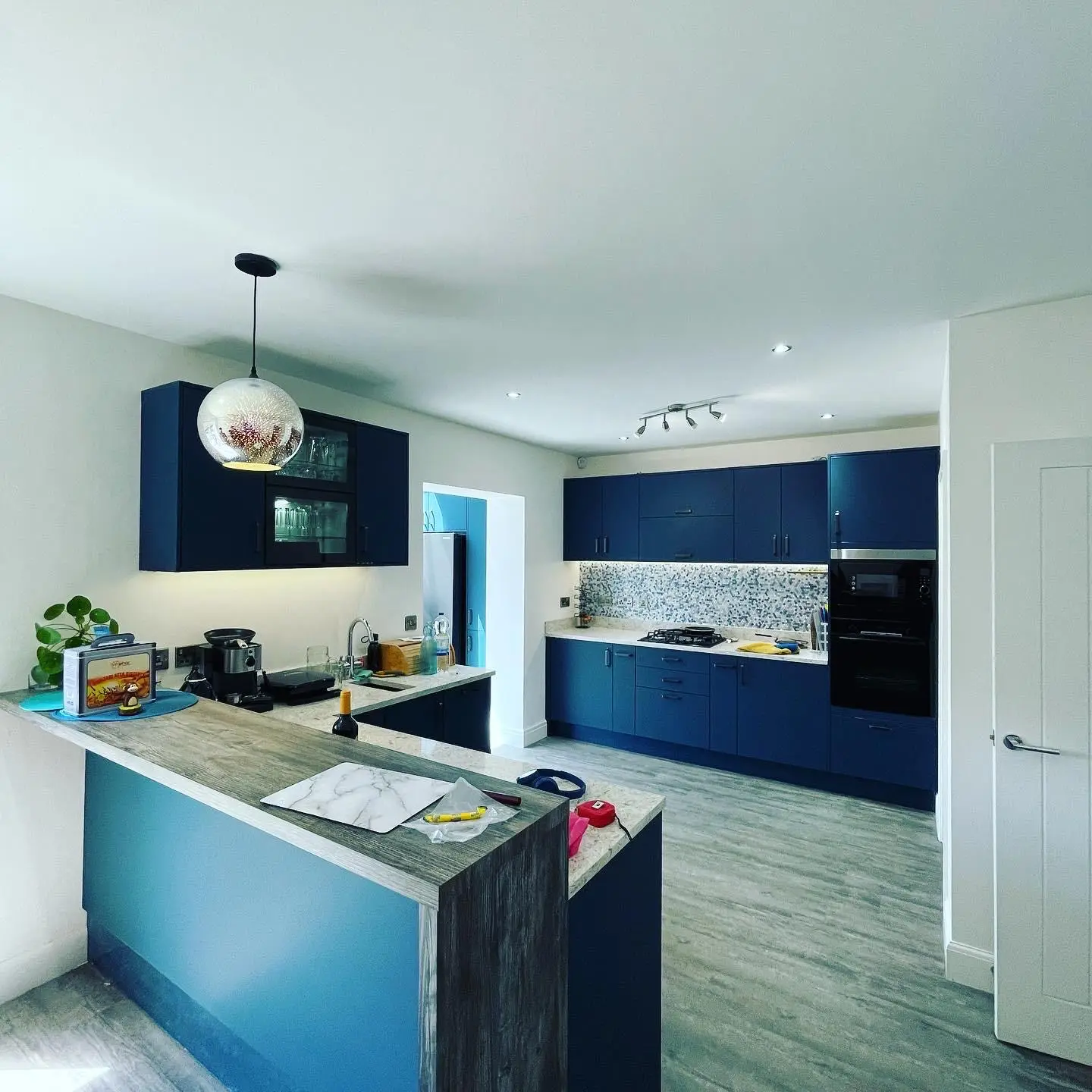 A blue kitchen with a globe pendant over the breakfast bar, a spotlight bar, downlights, LED tape under the wall units and a lit glass-fronted cabinet.