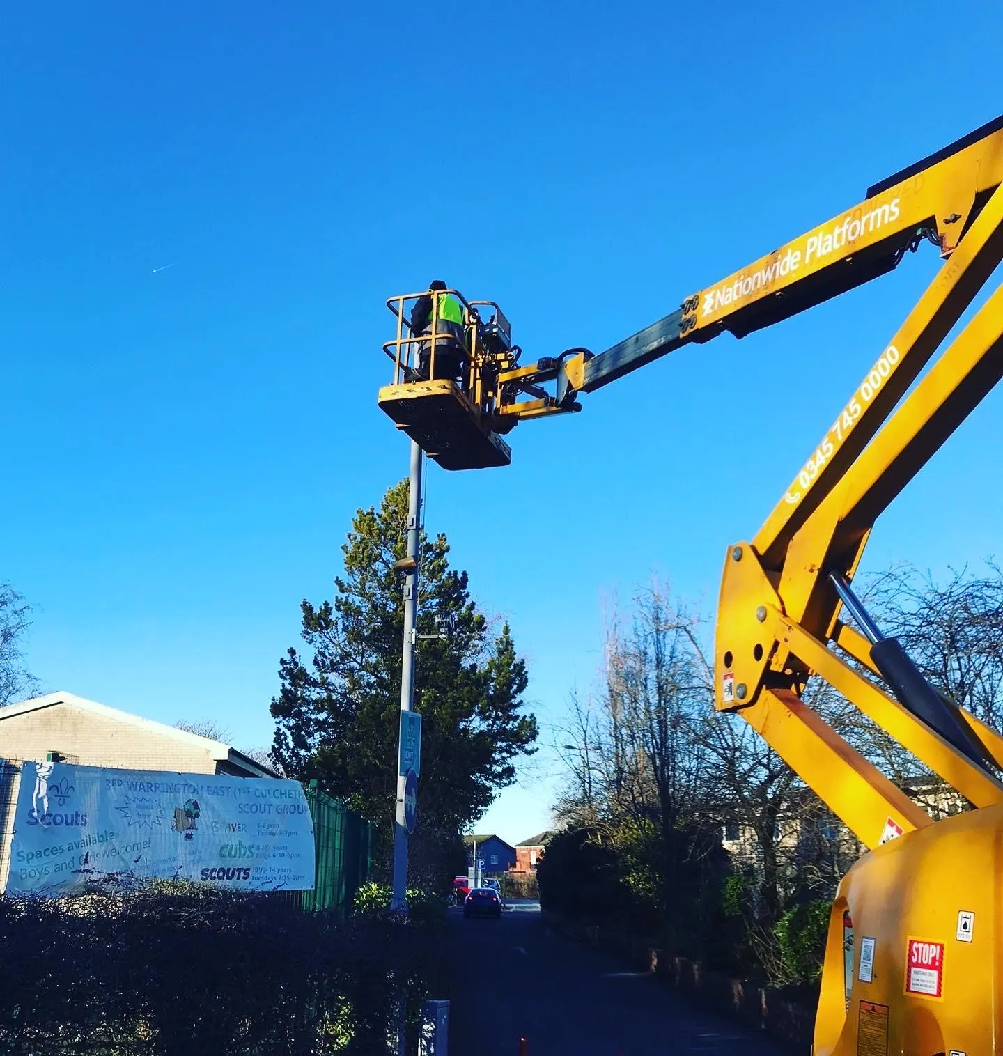 An electrician in hi-vis working at the top of a street lighting column from a boom lift, on an access road under a blue sky.