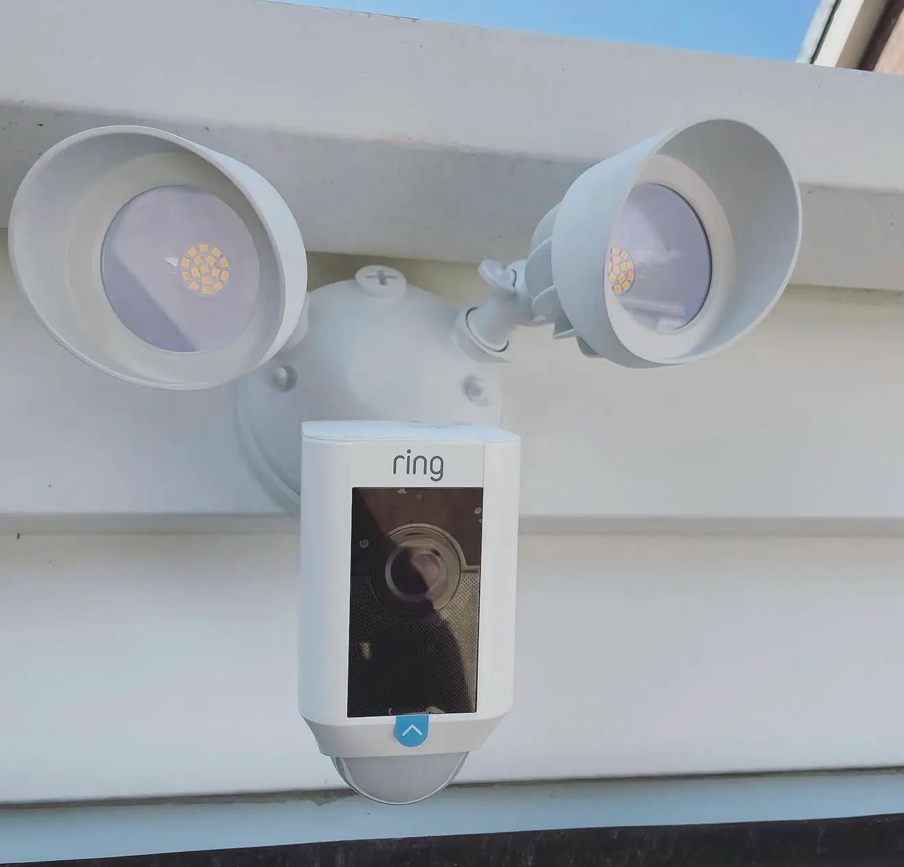 A white Ring floodlight camera with twin LED lamps and a motion sensor, fitted to a white fascia board.