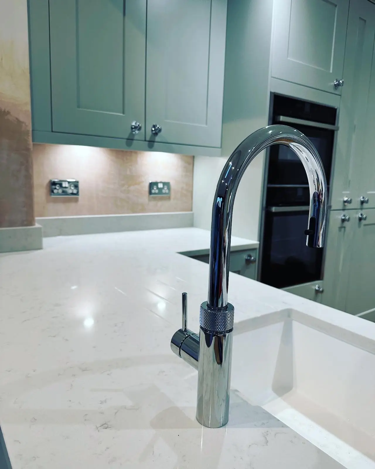 Under-unit lighting over a plastered splashback fitted with two polished chrome double sockets, seen past a chrome tap, with built-in ovens beyond.