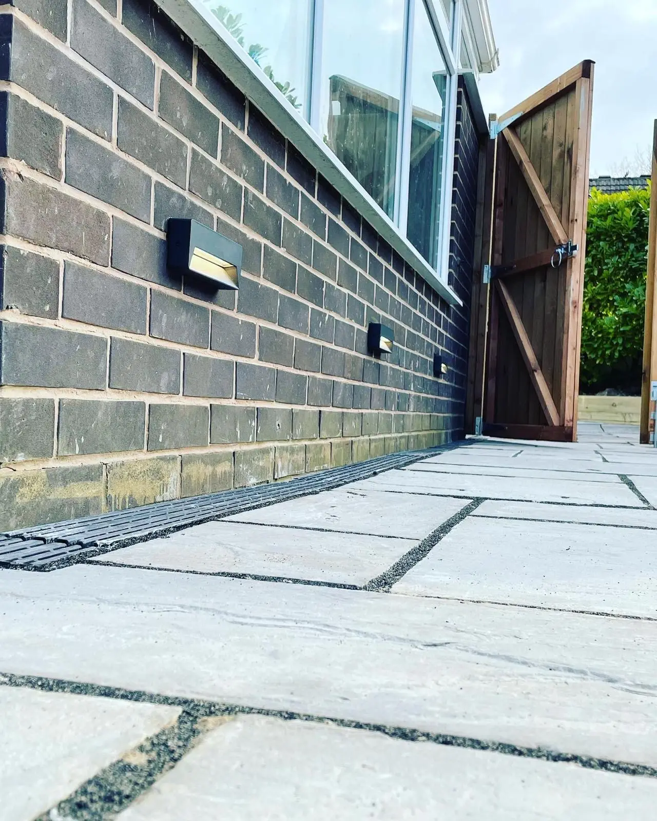 Three low-level black brick lights set along the wall of a side passage, beside an open timber gate.