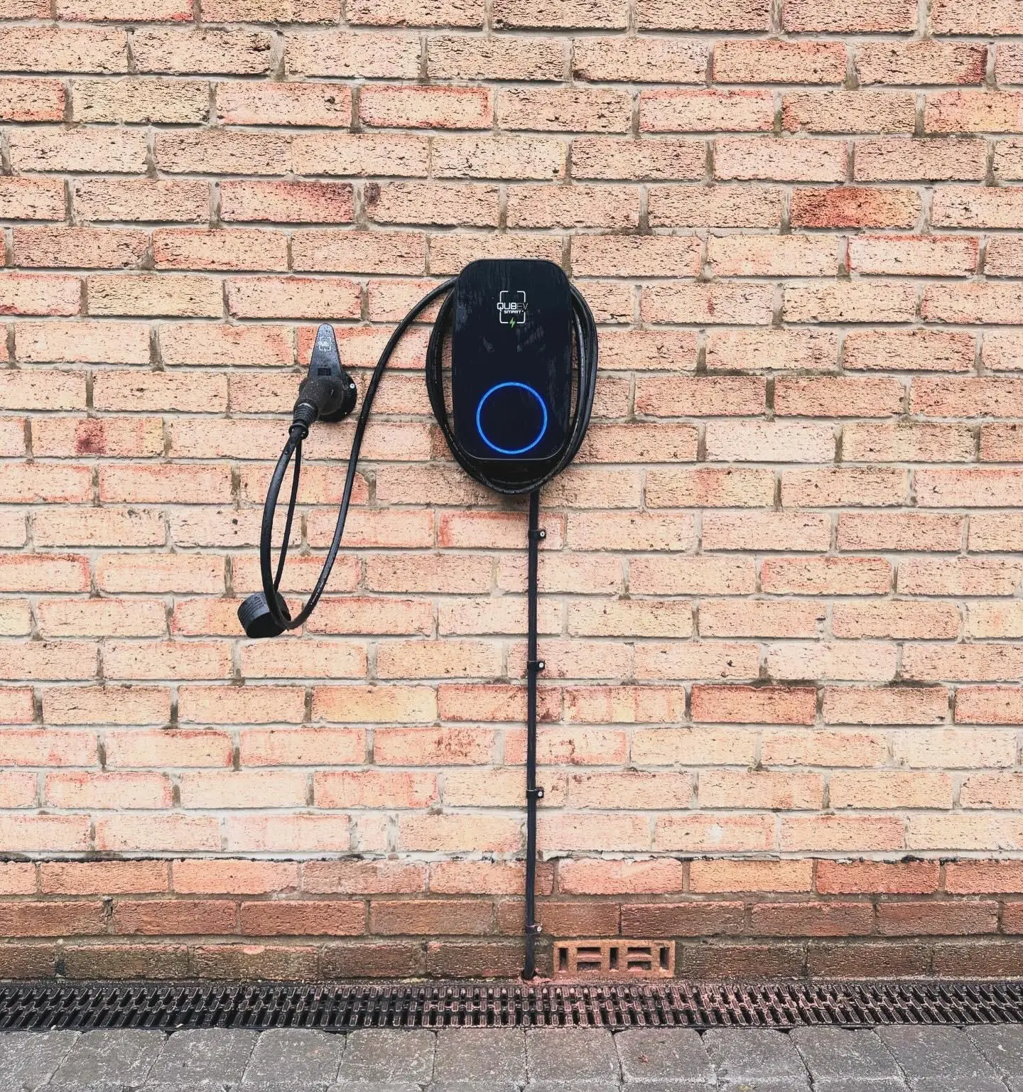 An EV charger on a brick wall seen from a lower angle, with its supply run in black conduit down to ground level.