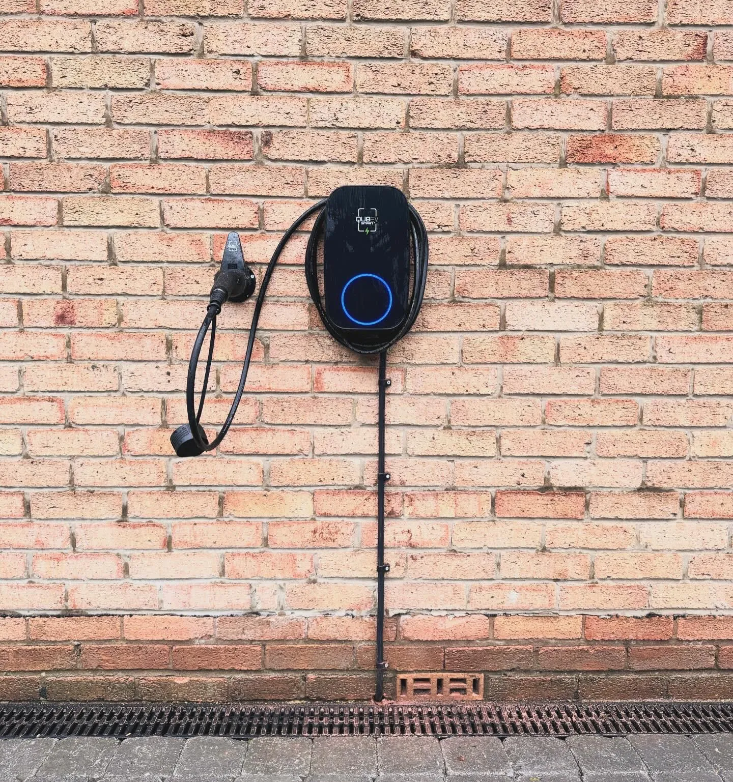An EV charger on a brick wall seen from a lower angle, with its supply run in black conduit down to ground level.