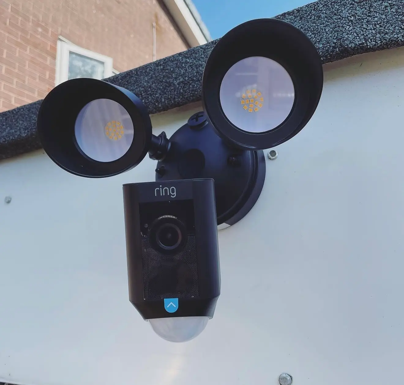 A black Ring floodlight camera with twin LED lamps and a motion sensor, fitted to a white fascia under the roof edge against a blue sky.