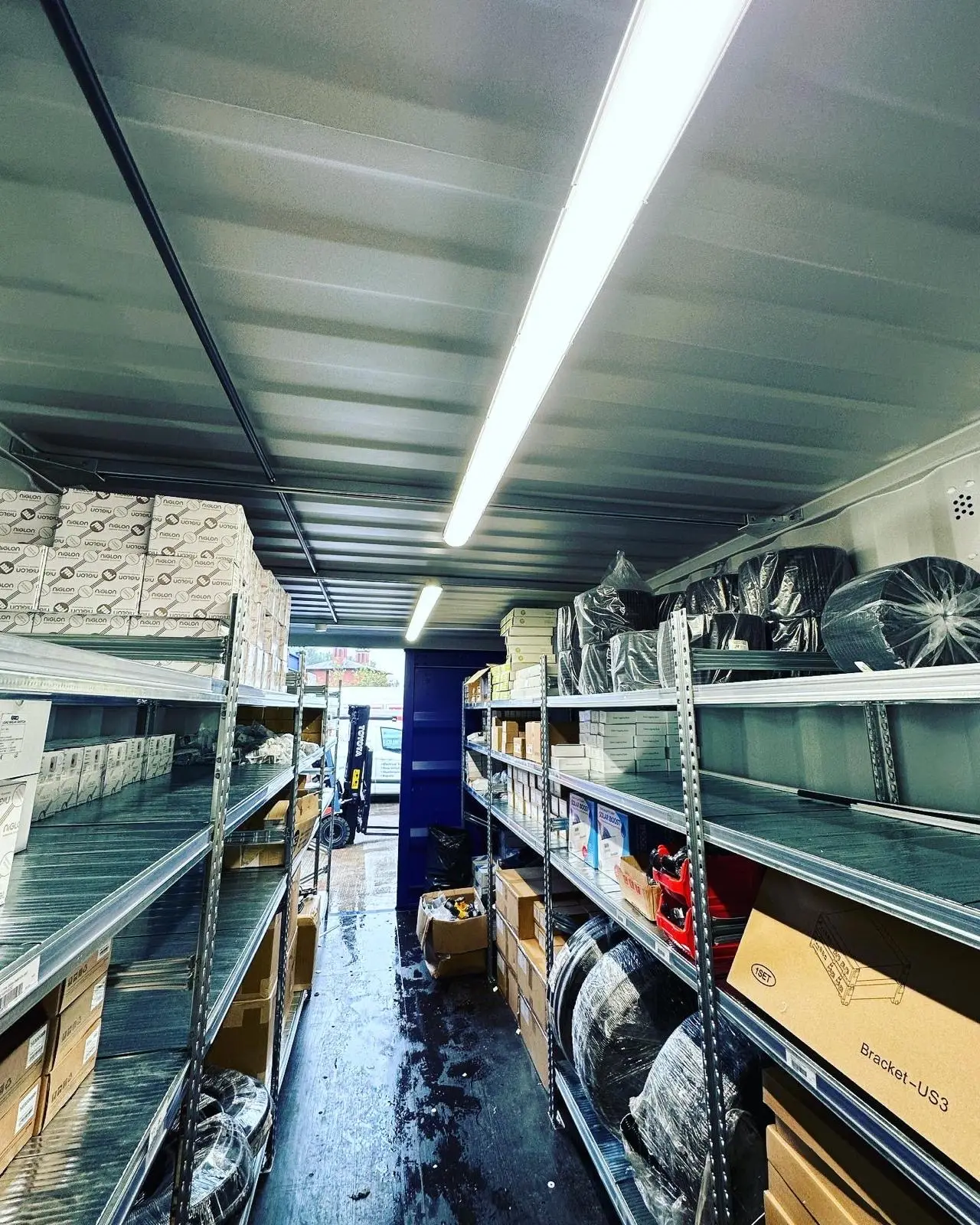 LED batten lights running along the ceiling of a storage container racked with stock on both sides.