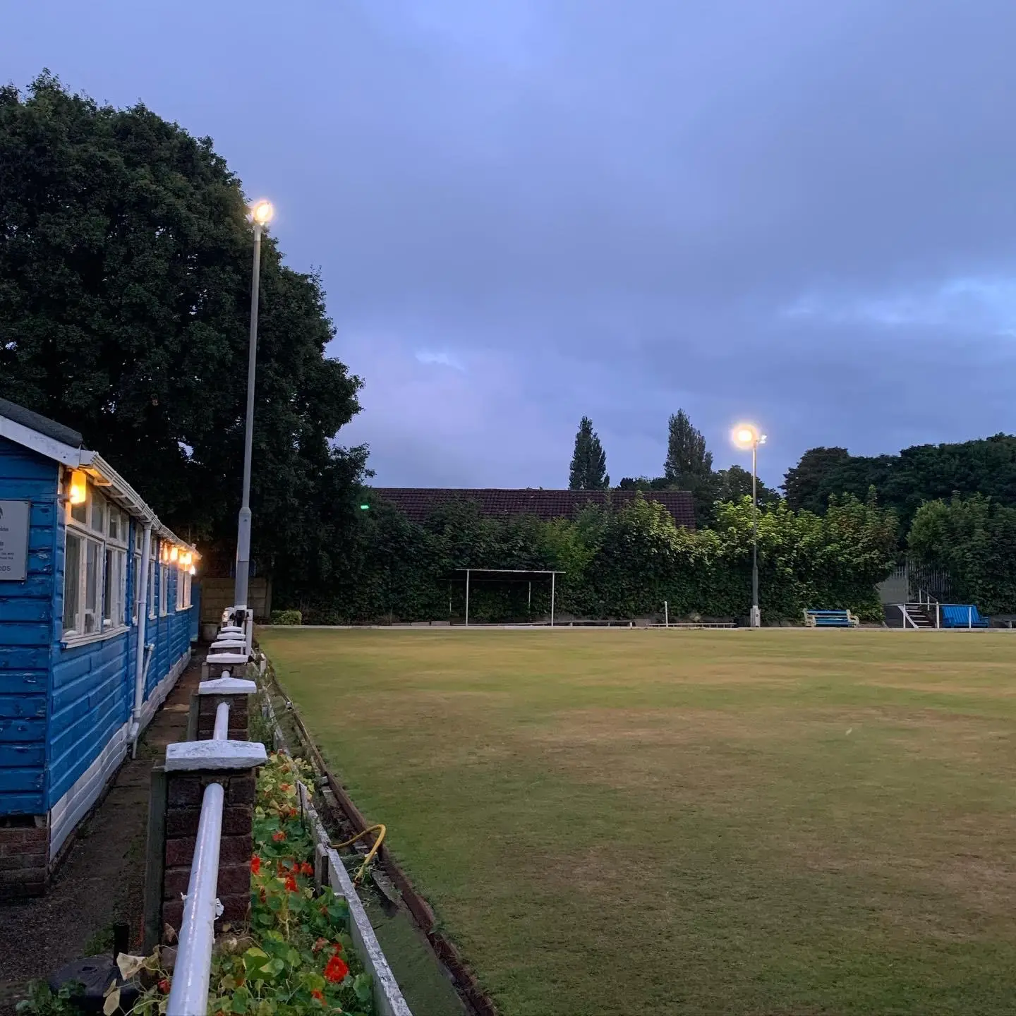 Two floodlights on columns lit at dusk beside a bowling green, with lights along the eaves of the timber clubhouse.