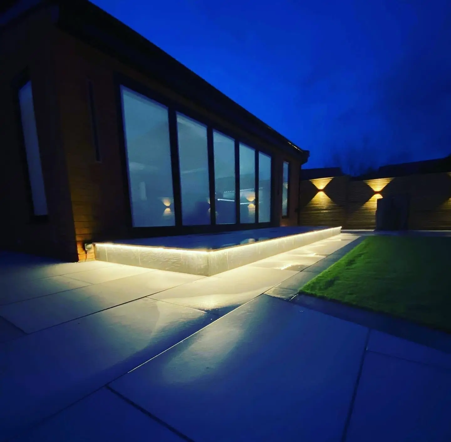 Close-up of LED tape lighting under a raised patio step at dusk, with up and down lights on the fence behind.