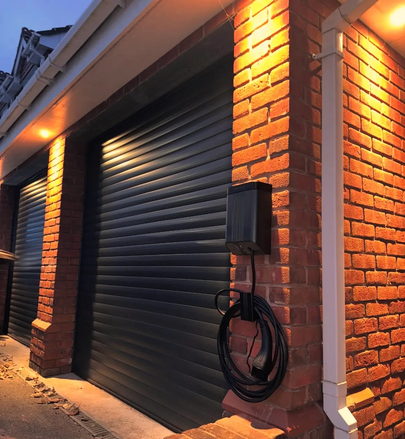 A black EV charger on a brick pillar between two garage roller doors, lit at dusk by soffit downlights.