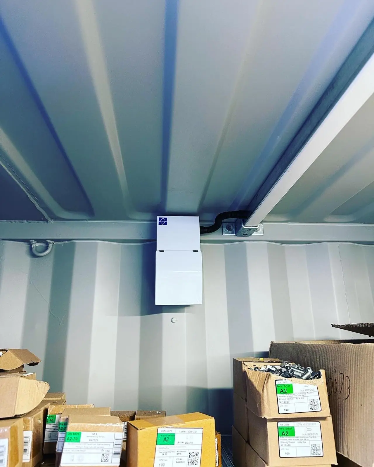 A small white consumer unit fixed to the top rail inside a storage container, fed in black flexible conduit, with a Unistrut bracket beside it.