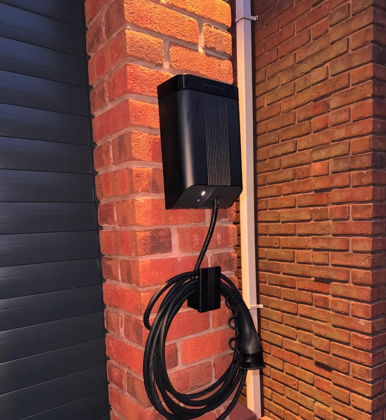 A black EV charger with its cable coiled on the holster, mounted on a brick pillar beside a garage door and downpipe.