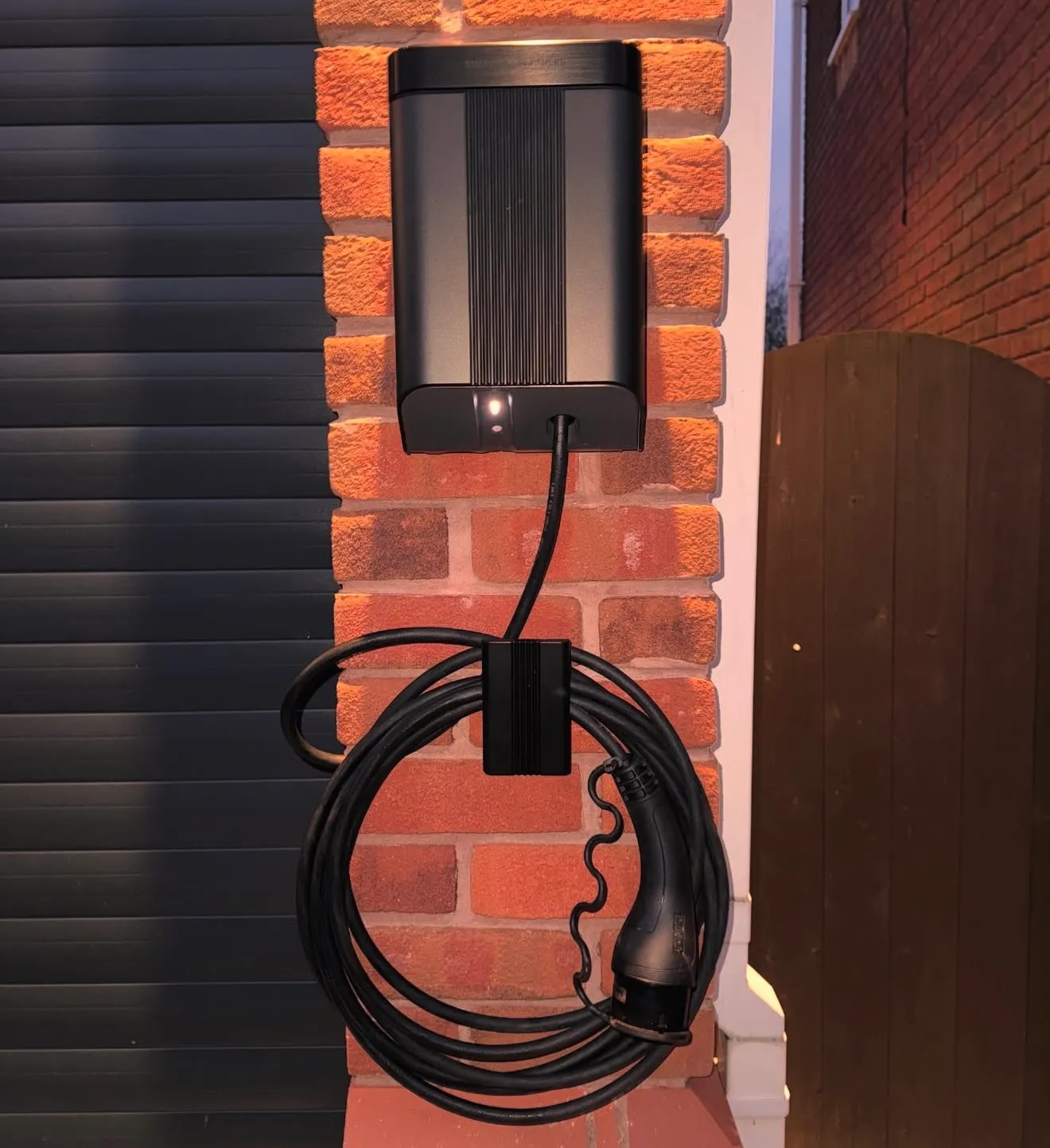 Front view of a wall-mounted EV charger on a brick pillar between a garage door and a timber gate, its charging cable coiled on a holster.