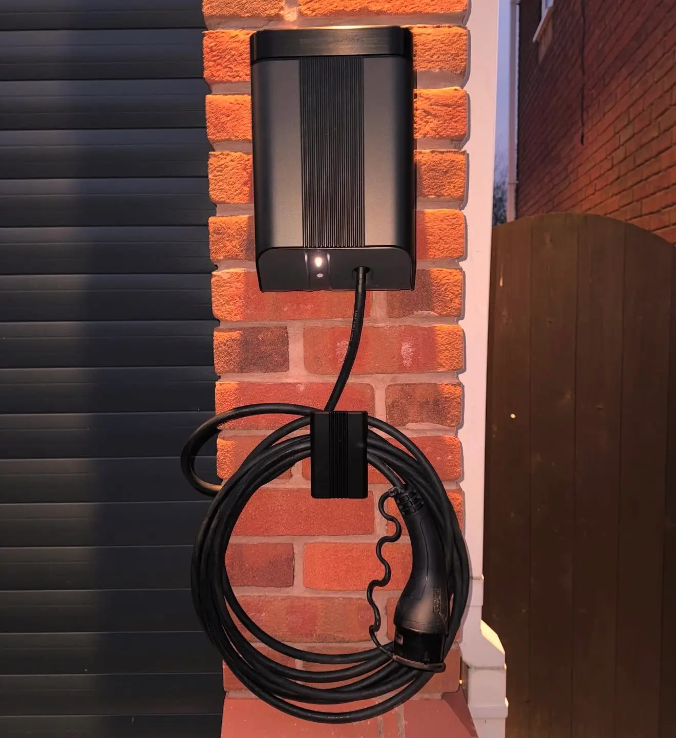 Front view of a wall-mounted EV charger on a brick pillar between a garage door and a timber gate, its charging cable coiled on a holster.