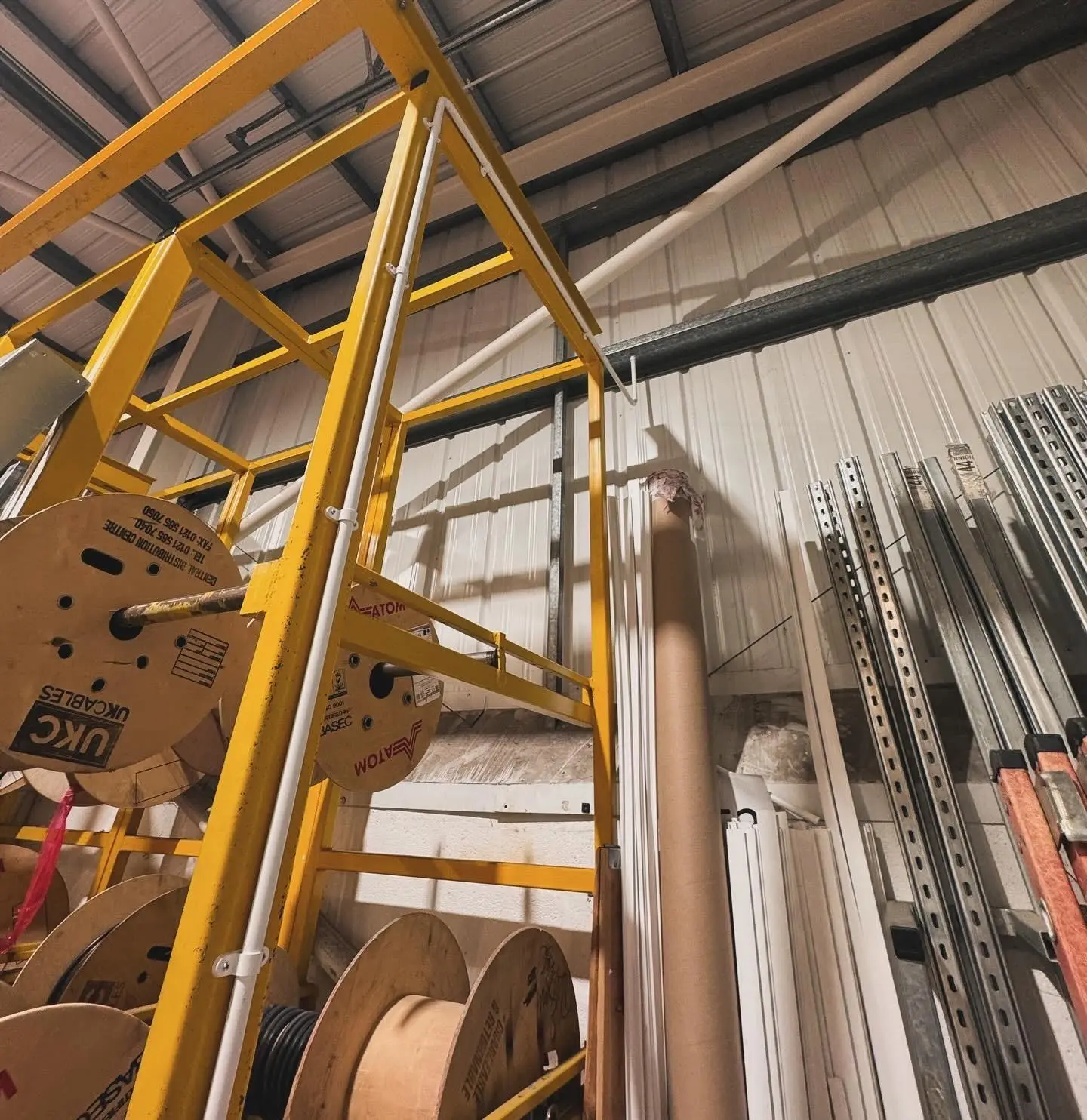 White PVC conduit run up a yellow cable drum rack and across to the roof steelwork in a warehouse.