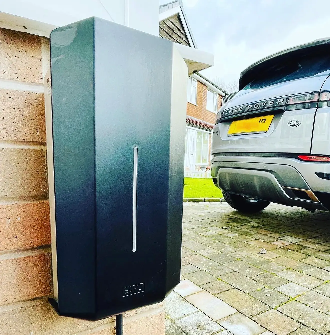 A dark grey GARO EV charger fixed to the end of a brick wall beside a block-paved driveway, with its supply in black conduit below.