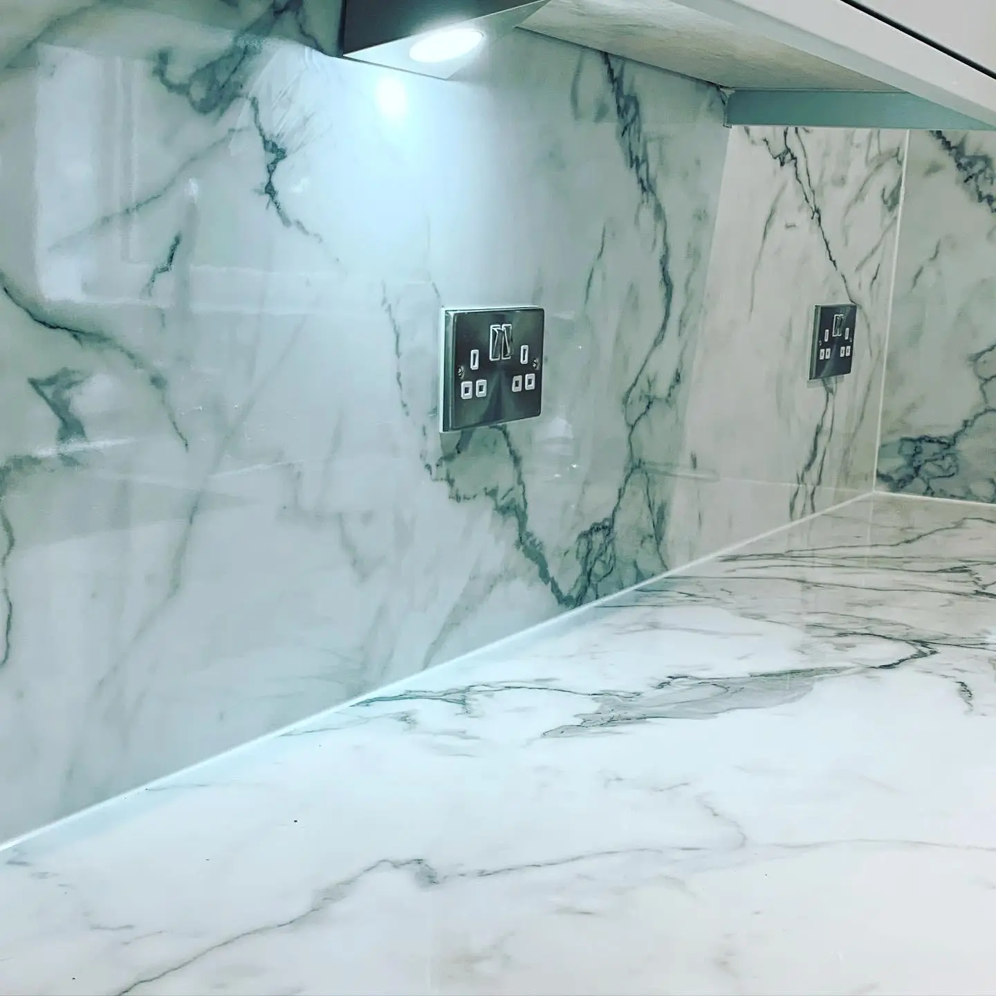 Polished chrome double switched sockets set into a marble-look splashback, lit by an LED under-cabinet light.