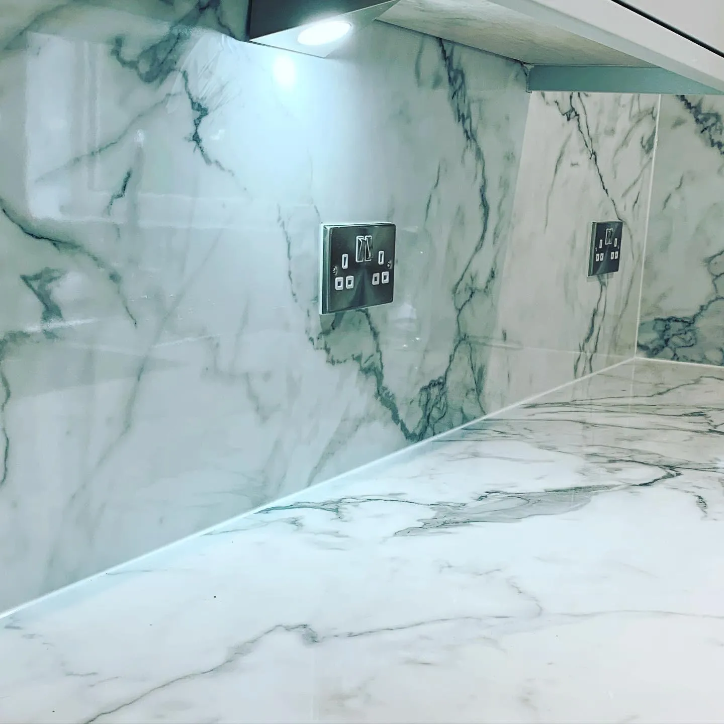 Polished chrome double switched sockets set into a marble-look splashback, lit by an LED under-cabinet light.