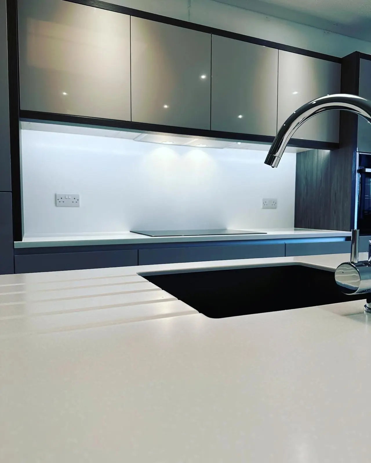 An extractor built in under the wall units with twin lights over an induction hob, with LED under-unit lighting and a white double socket either side, seen across an island sink.