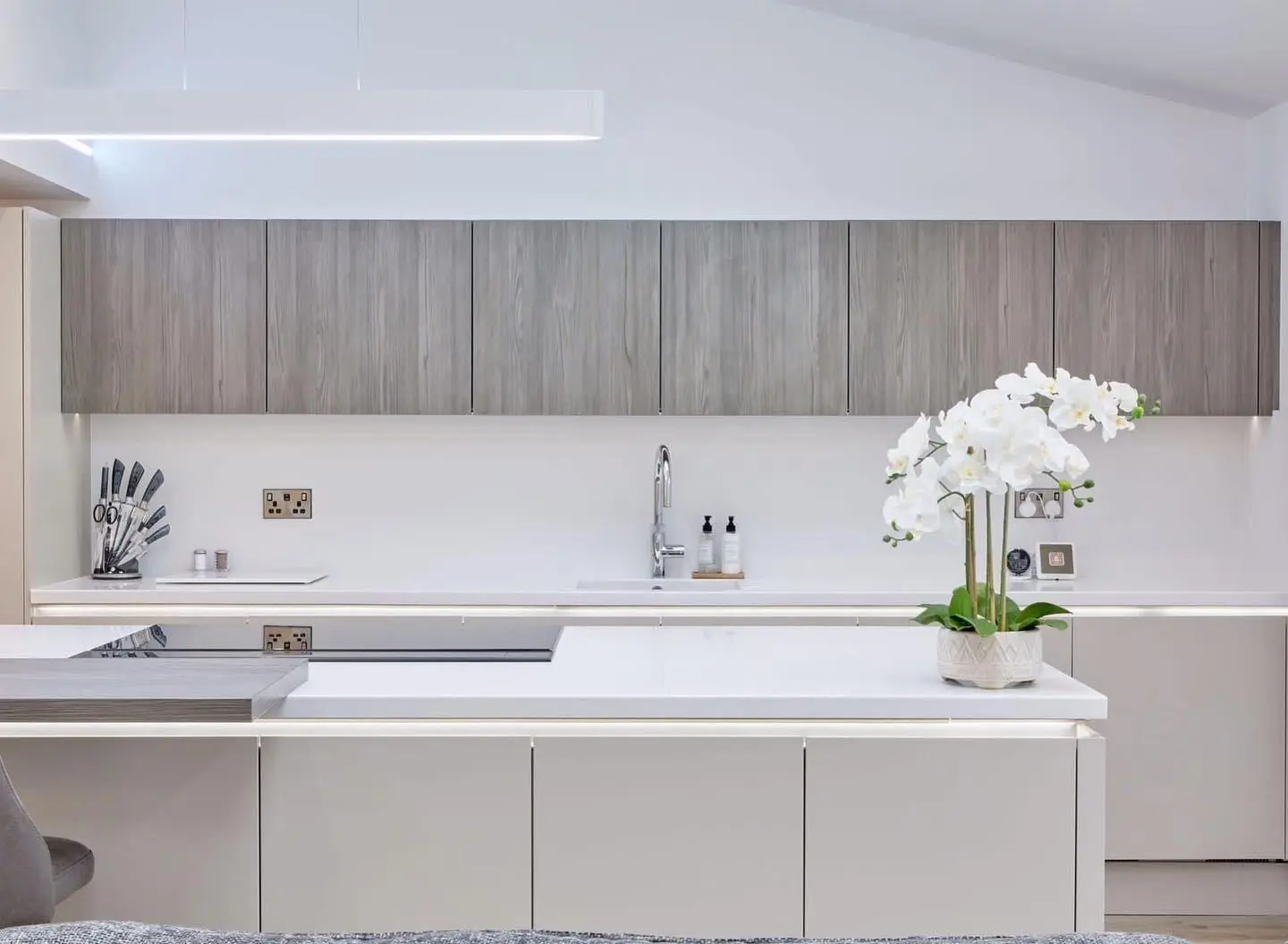 A linear LED pendant above a handleless kitchen, with LED tape under the wall units and along the worktop edges, and brushed double sockets on the splashback.