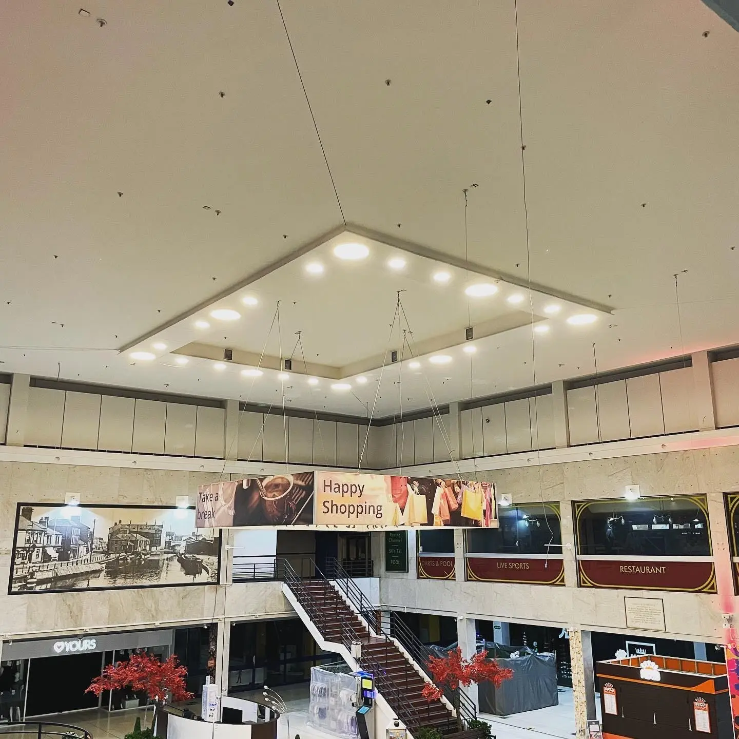 The suspended ceiling raft of LED downlights above a shopping centre atrium, seen from the upper floor over the stairs.