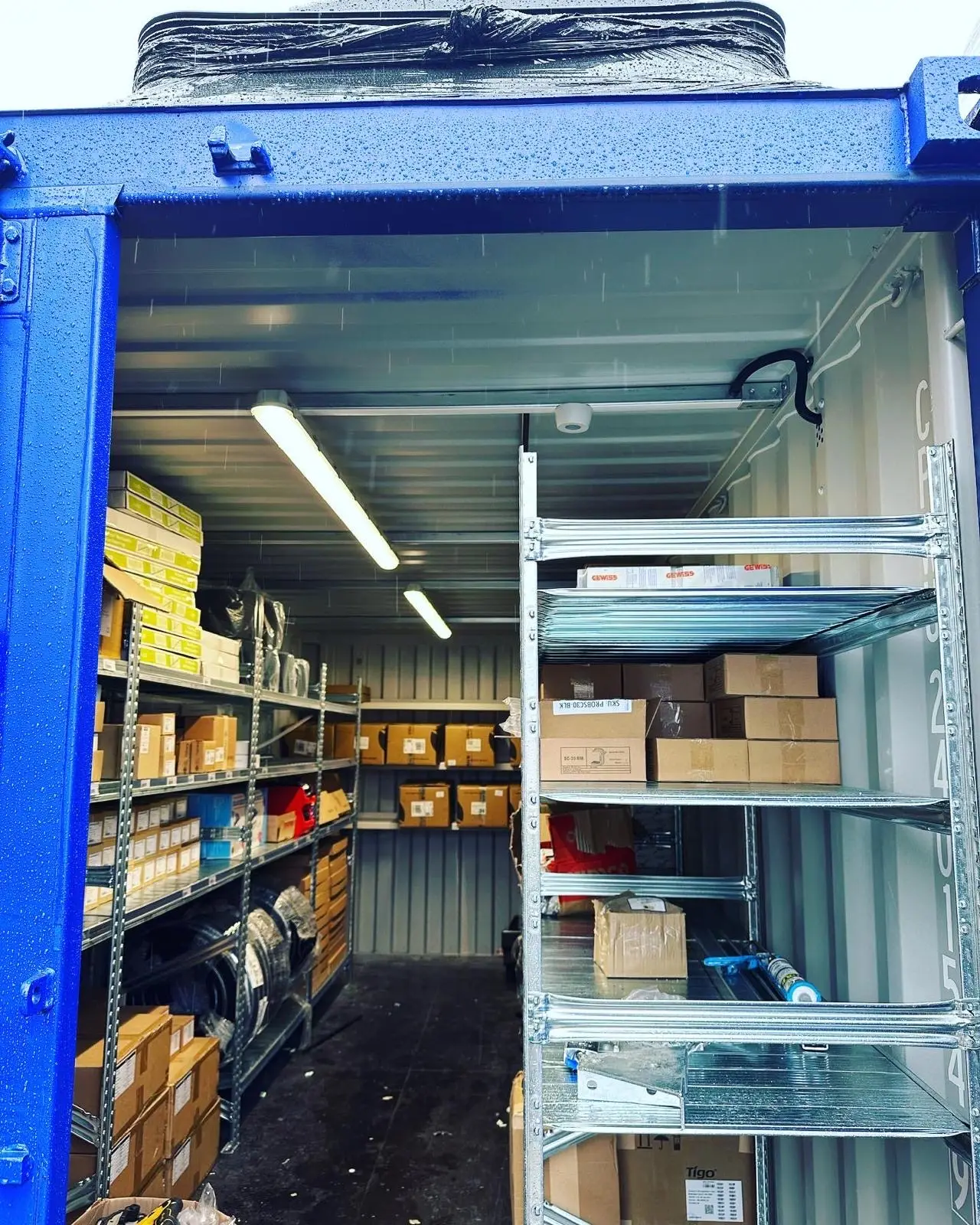 The open end of a storage container fitted with LED batten lights on the ceiling, fed in black flexible conduit along the top rail, above racked shelving.