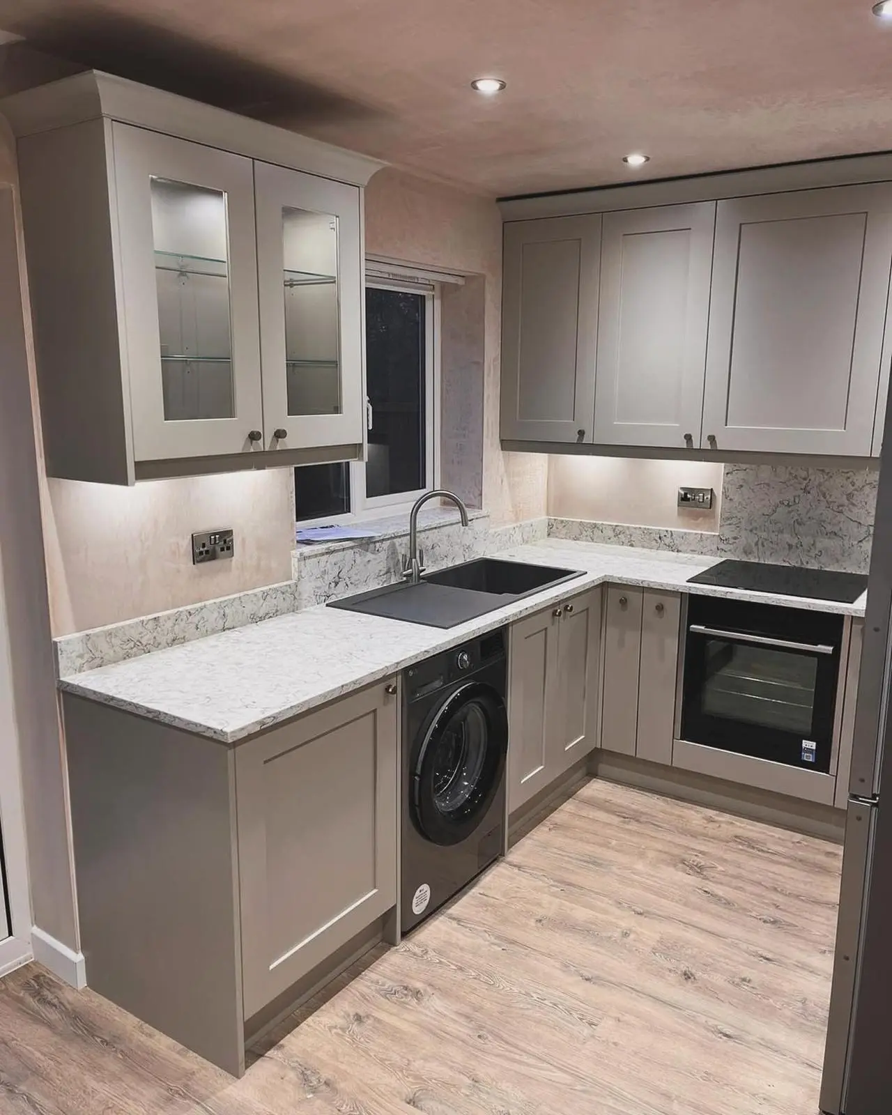 A grey shaker kitchen with downlights, lighting inside the glass-fronted wall cabinets, under-unit lights and brushed chrome sockets either side of the window.