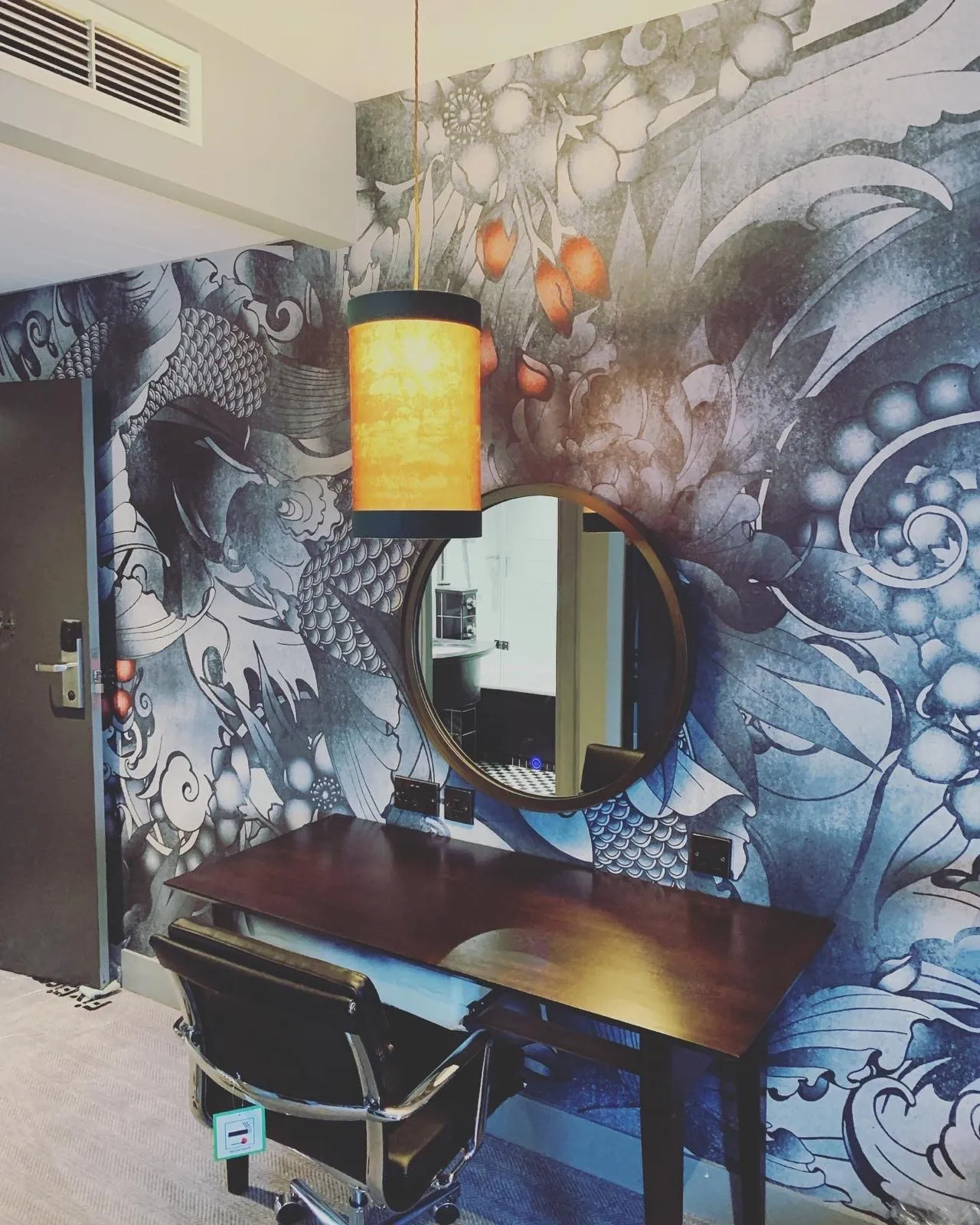 A hotel room desk area with a fabric drum pendant over the desk and dark-finish sockets set into the wall at desk height.