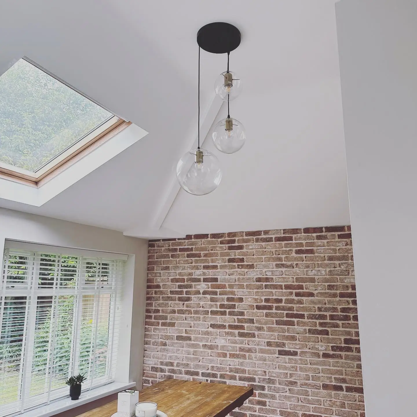 A triple cluster pendant of clear glass globes on a black ceiling plate, hung from a vaulted ceiling beside a rooflight over a dining table.