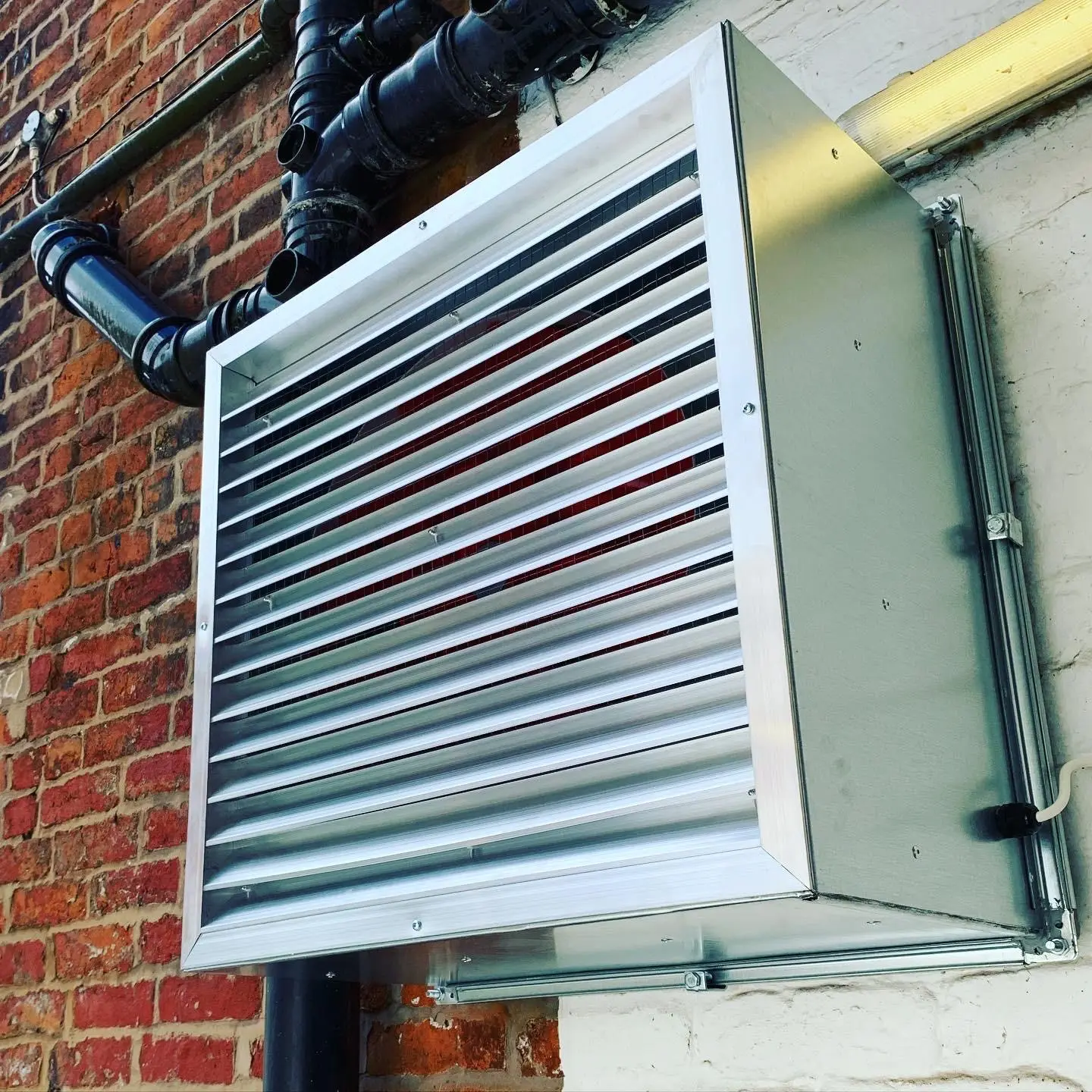 A galvanised industrial fan heater mounted on a Unistrut frame on a painted brick wall, its supply cable entering through a gland.