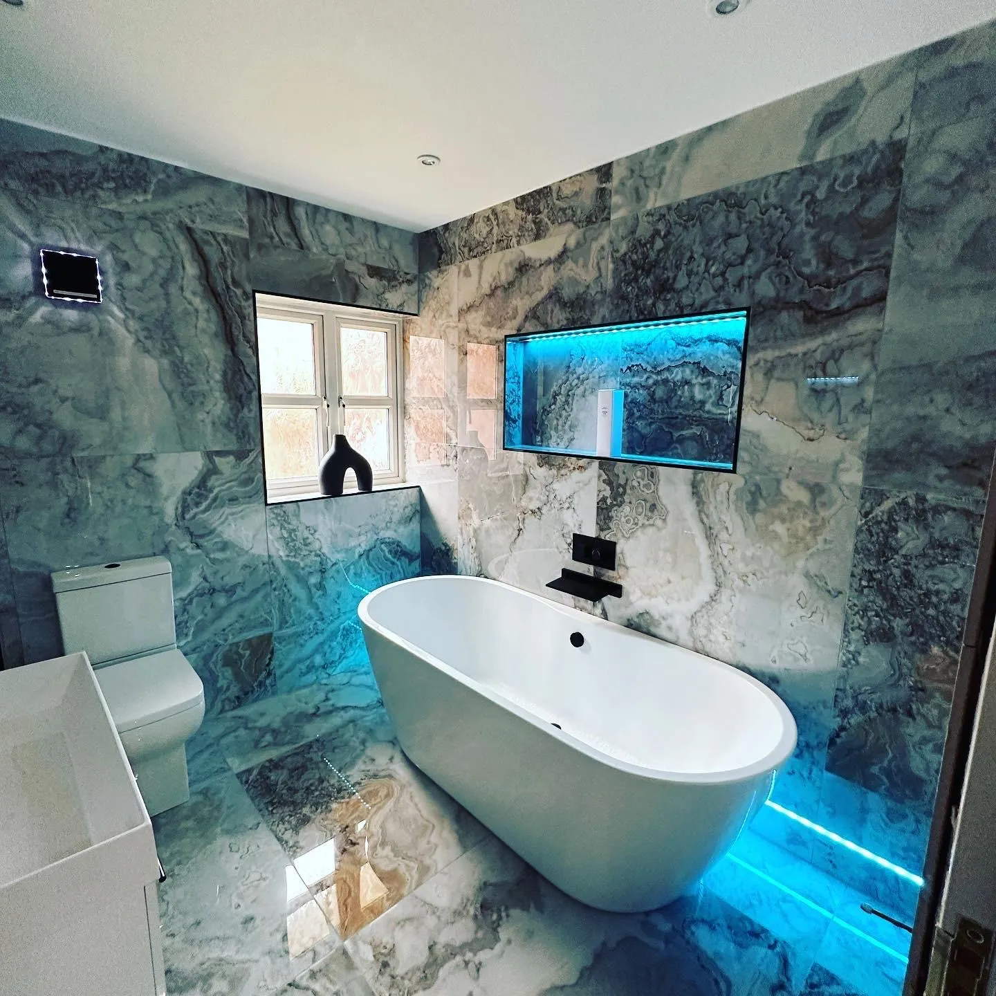 A marble-tiled bathroom with blue LED tape recessed into a tiled niche above the freestanding bath and along a floor-level recess, plus downlights and a square wall extractor fan.