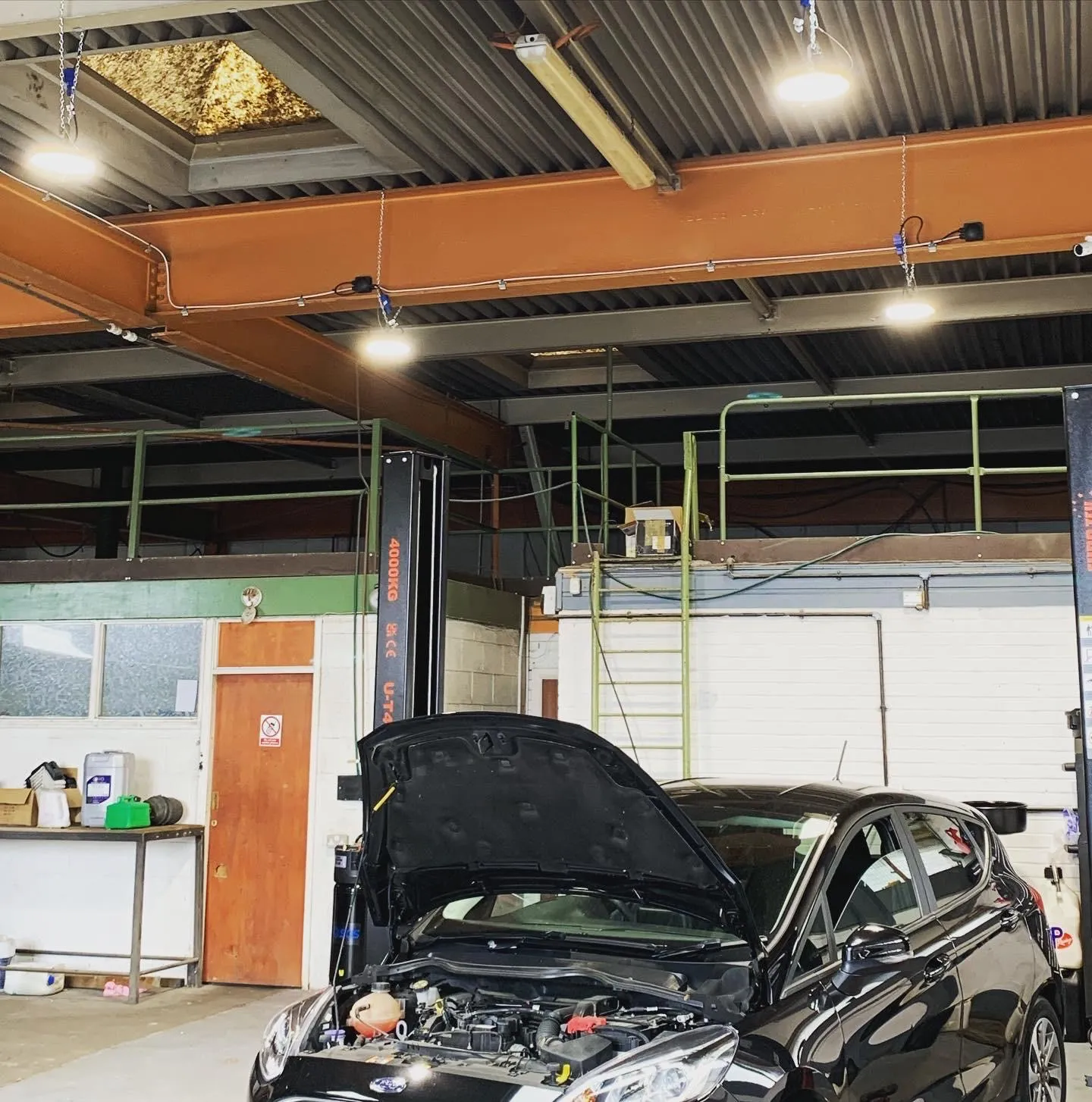 Round LED high bay lights hung on chains from the steel beams of a vehicle workshop, with their cabling clipped along the beams.