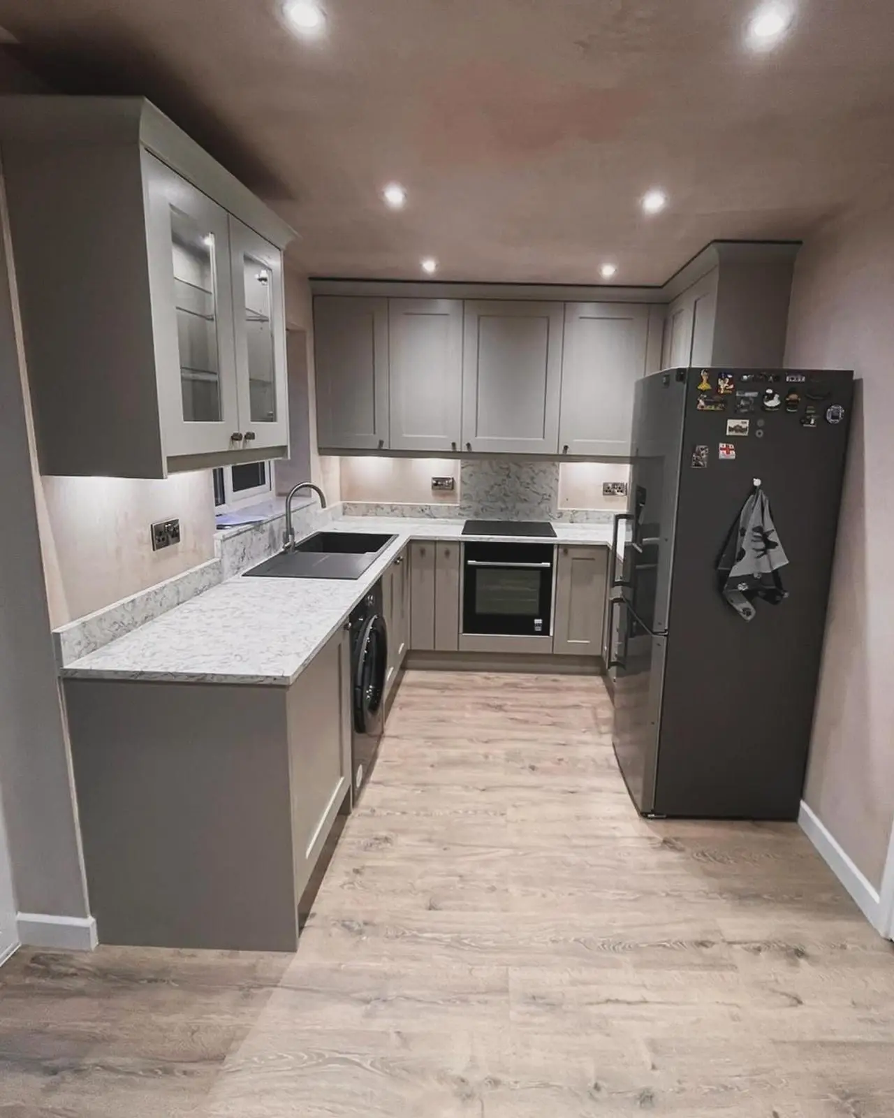 A grey shaker kitchen seen from the doorway, with LED downlights across the ceiling, under-unit lighting, a lit glass-fronted cabinet and brushed chrome sockets.