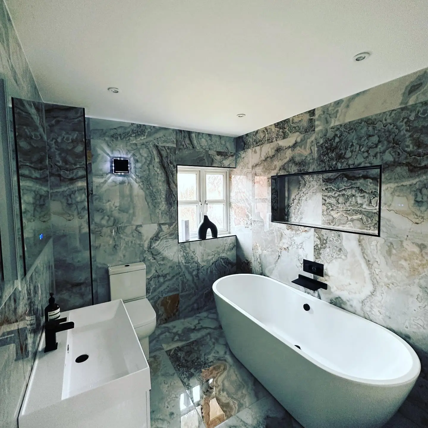 A marble-tiled bathroom with downlights, a lit square wall extractor fan and the edge of an LED mirror with a touch sensor, beside the freestanding bath.