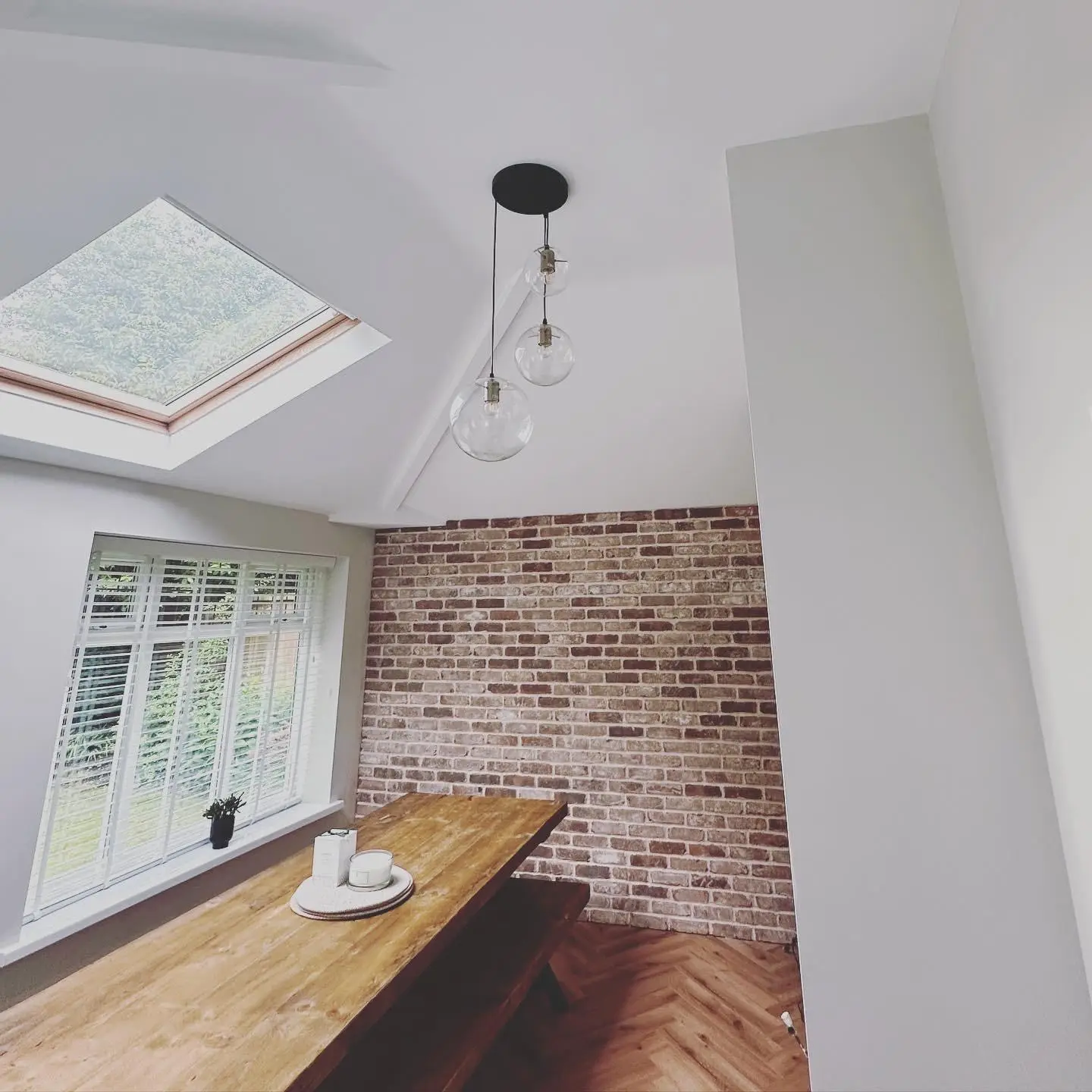A wider view of the triple glass globe pendant hanging from the vaulted ceiling over a dining table, beside a rooflight and a brick slip wall.