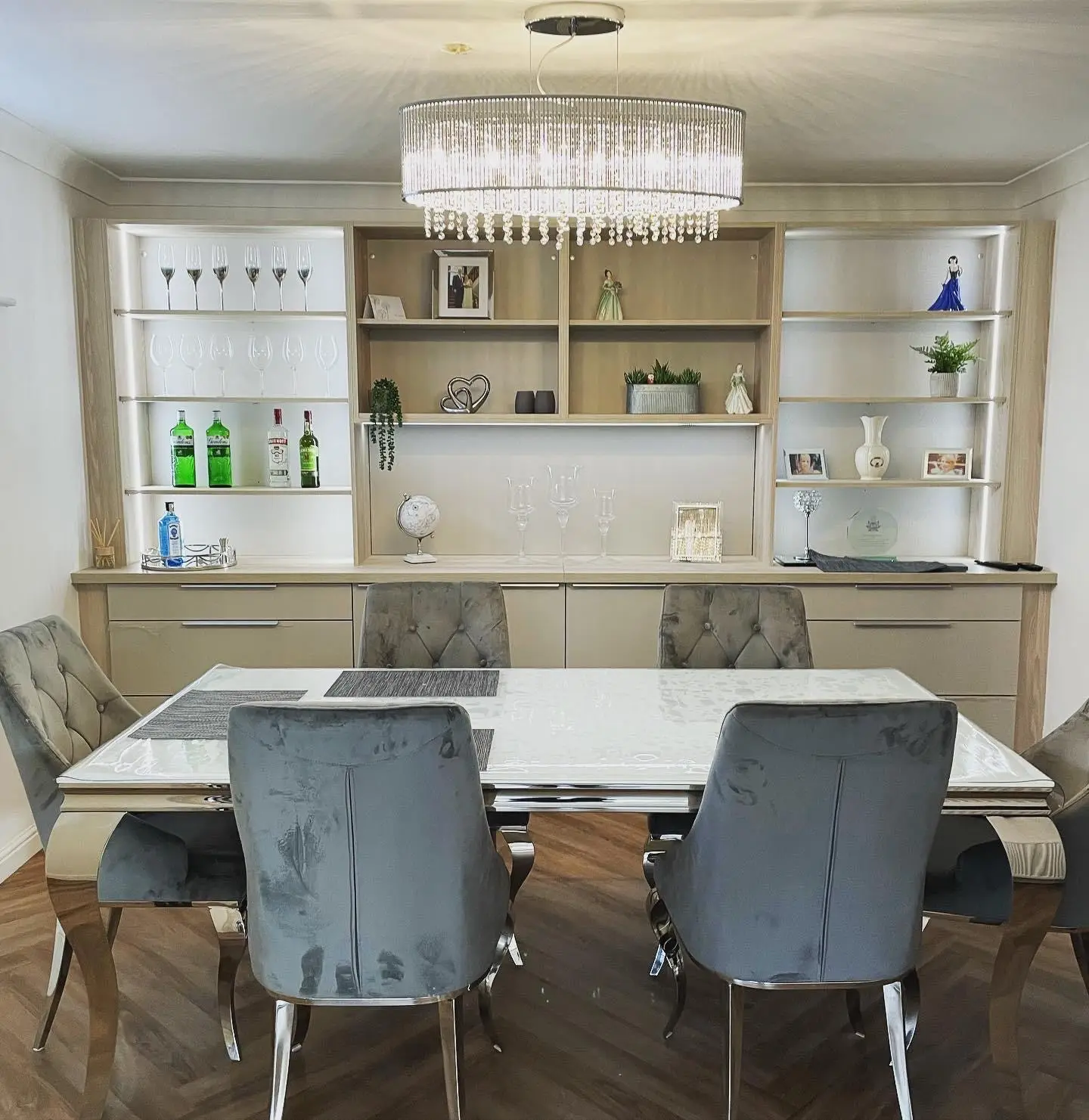 A large crystal drum chandelier over a dining table, in front of built-in display shelving lit with LED tape.