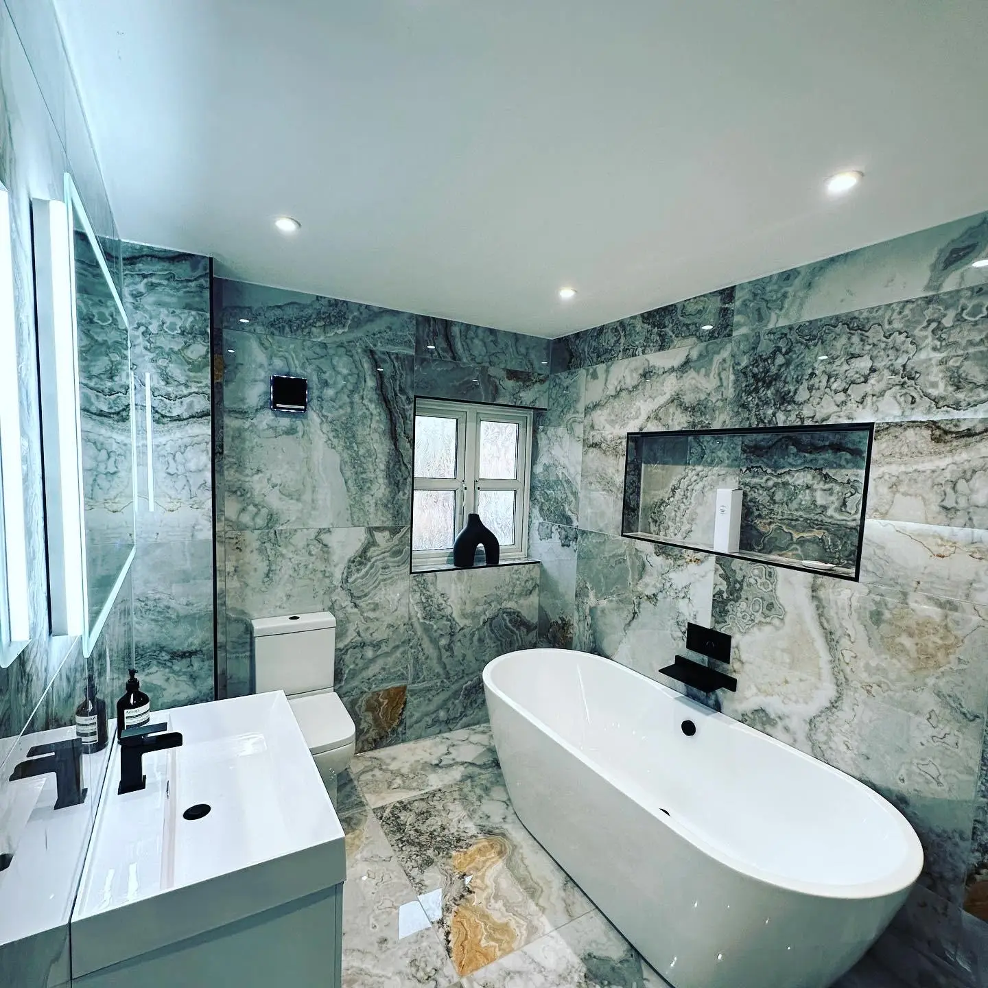 A marble-tiled bathroom with an LED mirror lit at its edges above the basin, downlights, a wall extractor fan and a recessed niche over the freestanding bath.