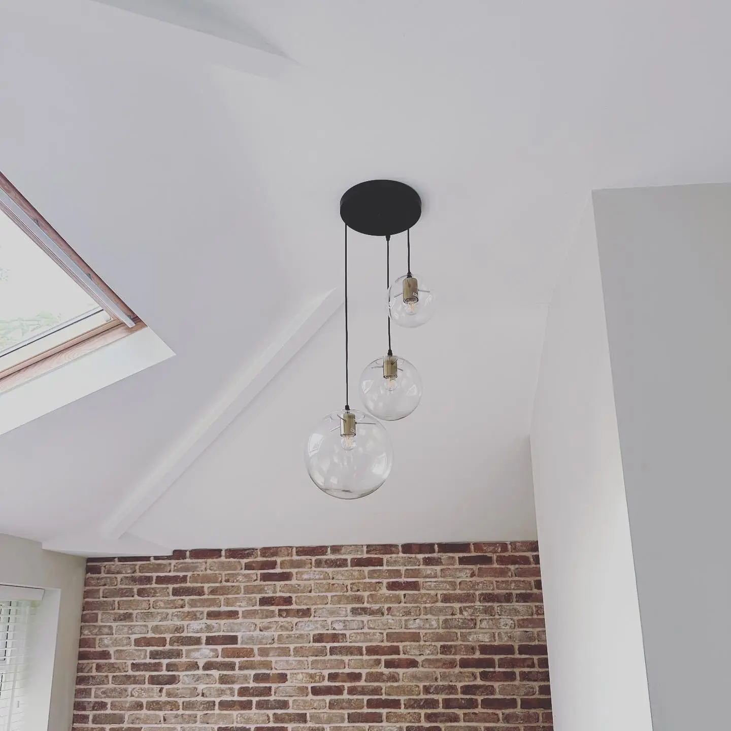 Close-up of a three-light cluster pendant with clear glass globes on a black ceiling plate, against a vaulted ceiling and brick slip wall.