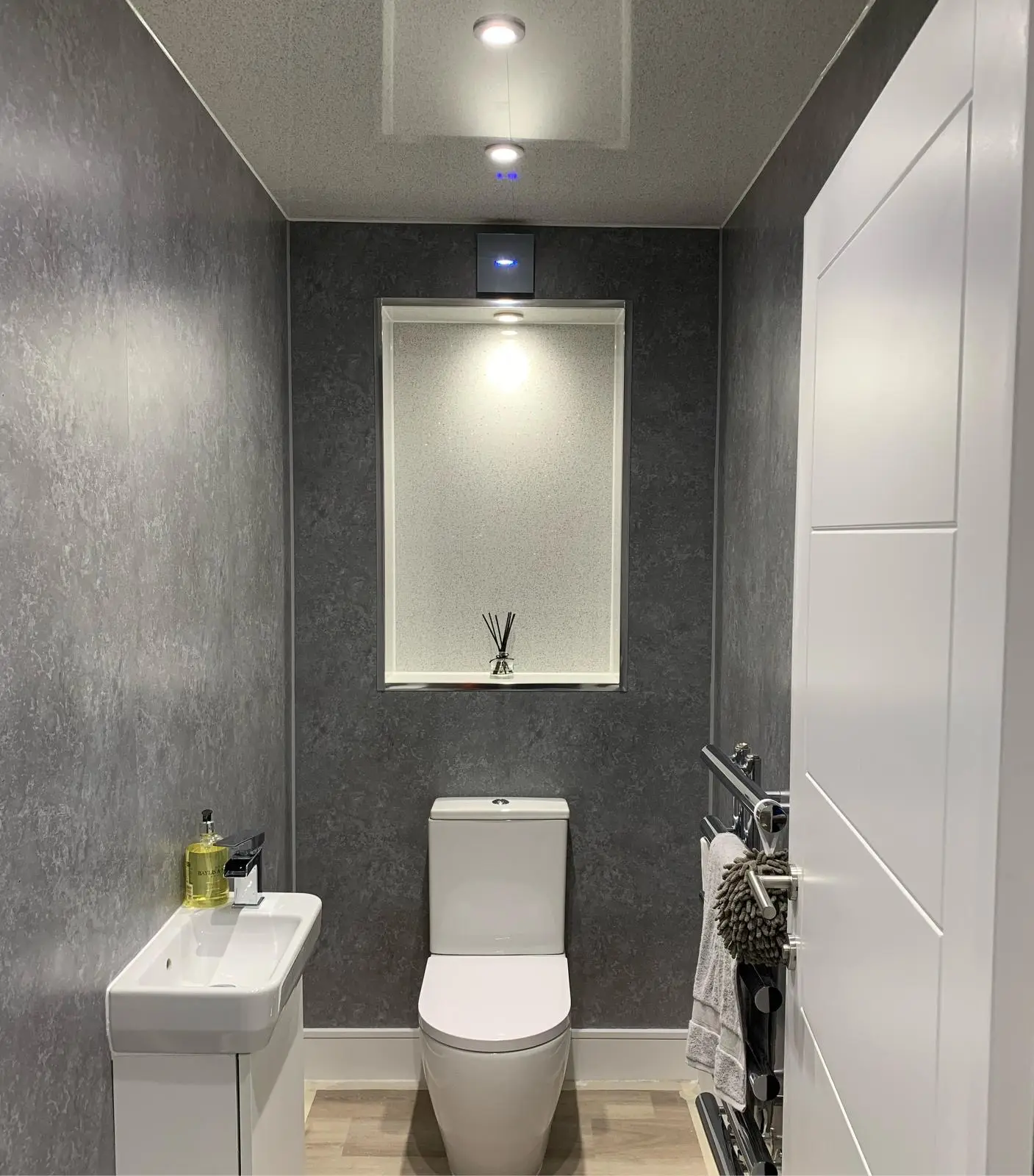 A cloakroom with two LED downlights, a black wall extractor fan with a blue indicator and a small downlight in the top of a recessed niche above the toilet.
