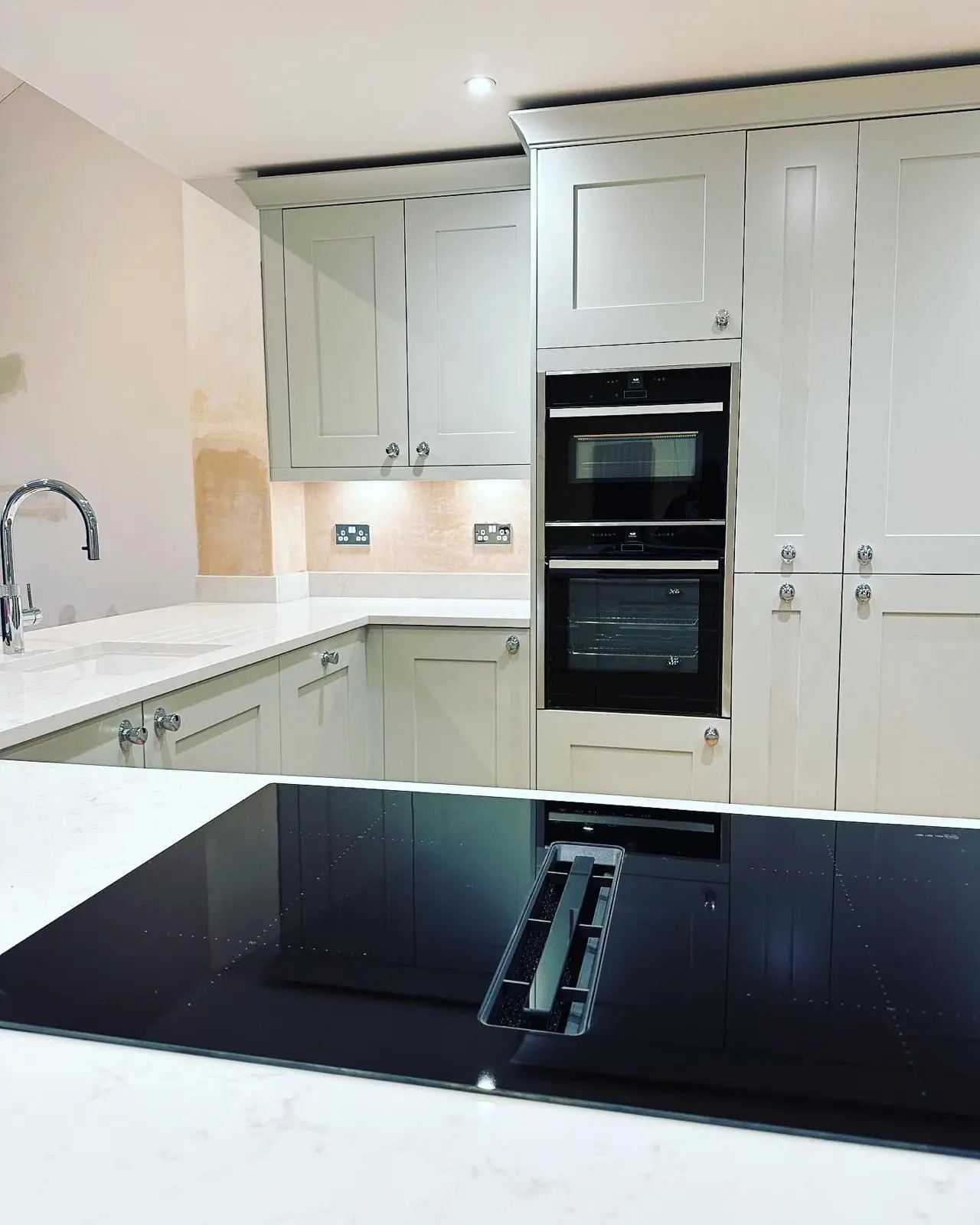 An induction hob with a built-in downdraft extractor in the foreground, stacked built-in ovens in a tall housing, and chrome double sockets under the wall-unit lighting.