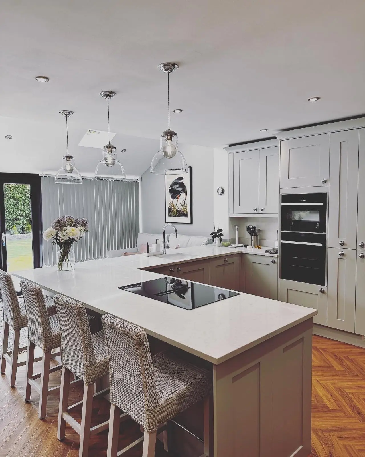 Three clear glass pendants and LED downlights over a peninsula breakfast bar with an induction hob, with stacked built-in ovens in the tall units.
