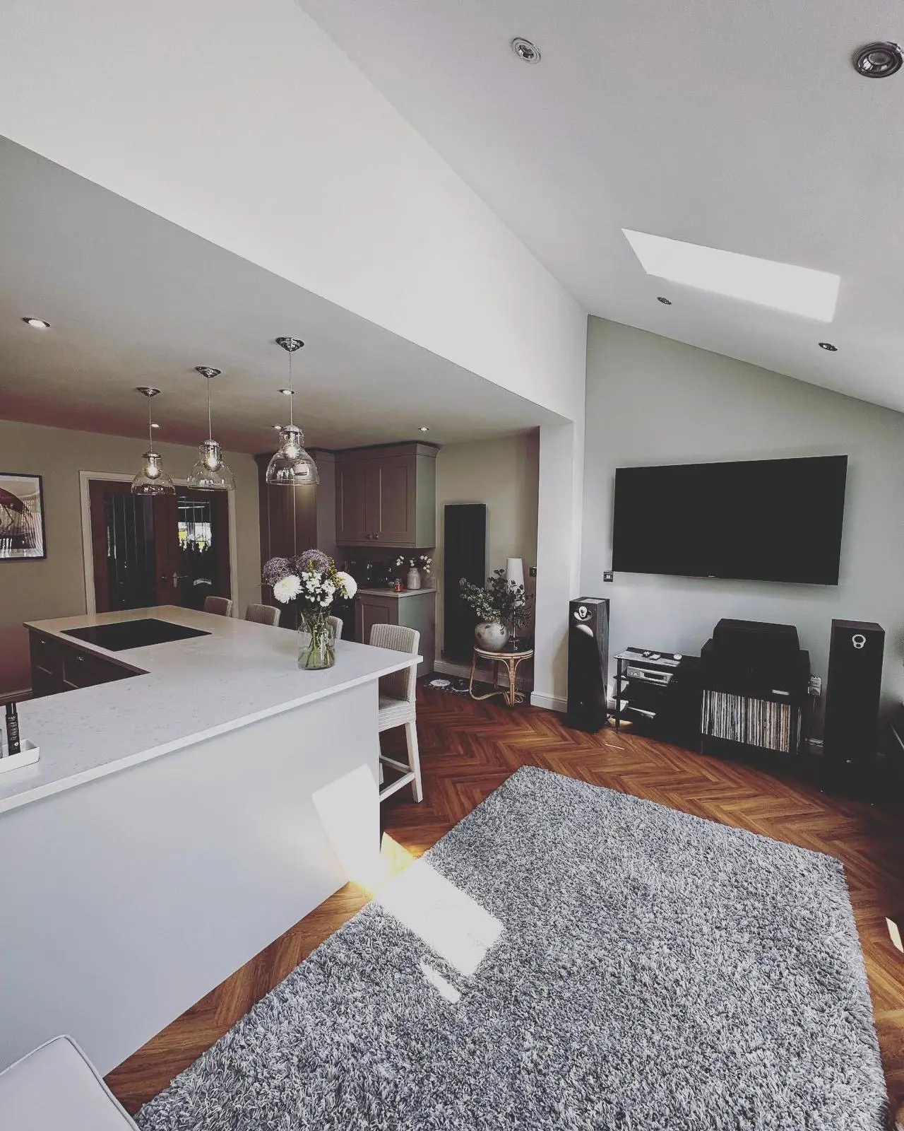 An open-plan extension with downlights in the vaulted ceiling, a wall-mounted TV and glass pendants over the peninsula beyond.