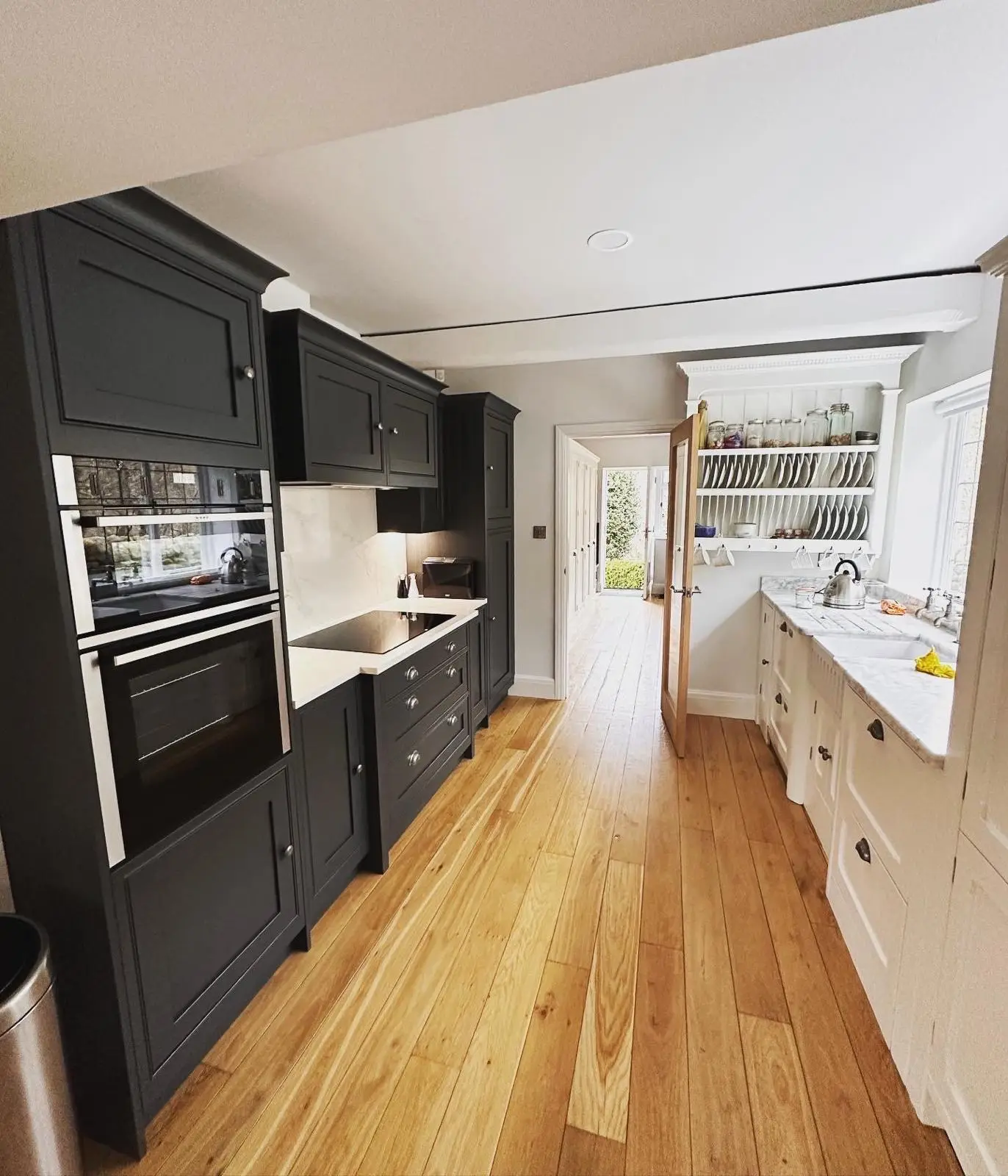 A dark shaker galley kitchen with stacked built-in ovens, an induction hob with lighting under the wall unit, and a downlight in the ceiling.
