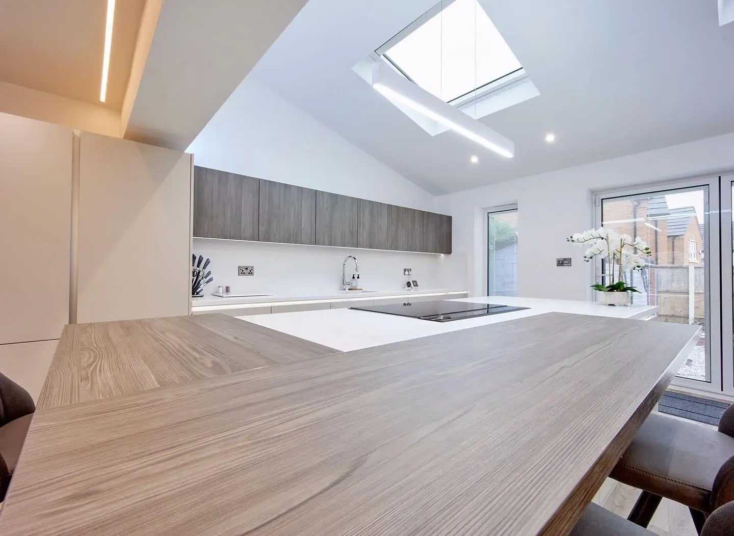 A linear LED pendant hanging beneath a rooflight over a kitchen island, with downlights, an LED profile in the ceiling bulkhead and LED tape under the wall units.