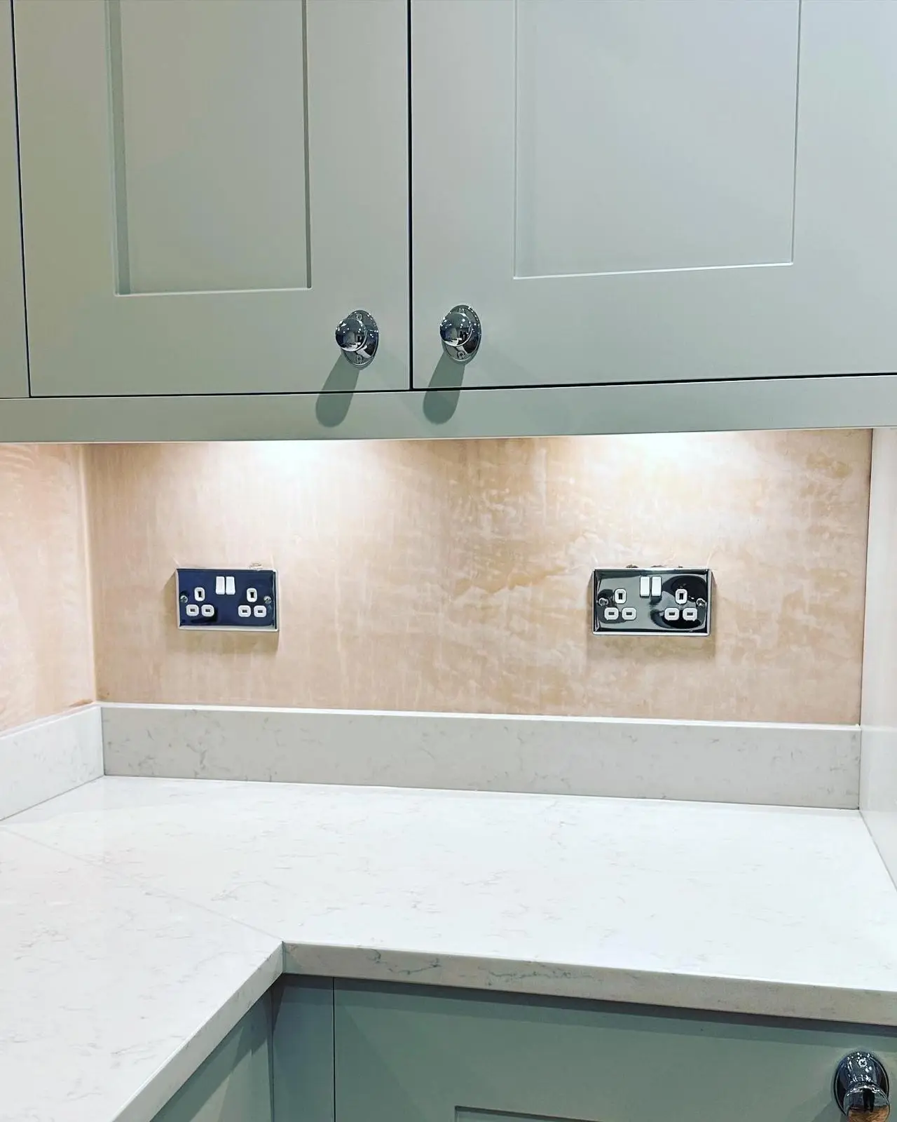 Two polished chrome double switched sockets with white inserts on a plastered splashback, lit by under-unit lighting.